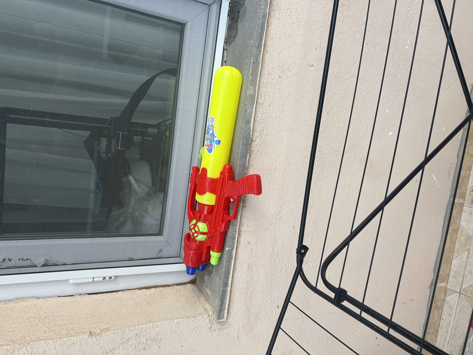 A bright yellow and red plastic water gun is mounted vertically on a wall next to a window. The water gun has a yellow cylindrical tank with a red handle and trigger mechanism. There are green and blue accents on the front of the gun. The gun has some text on the yellow tank which appears to be a logo and some smaller writing. The window is screened and slightly dirty, and appears to be part of a building. The wall is painted a light beige color and has some texture. There is a black metal grate or railing to the right of the water gun. The scene is outdoors, and the lighting suggests it is daytime, with no discernible weather conditions. There are no people visible in the image. The overall setting appears to be an exterior wall of a building, possibly in a residential area.