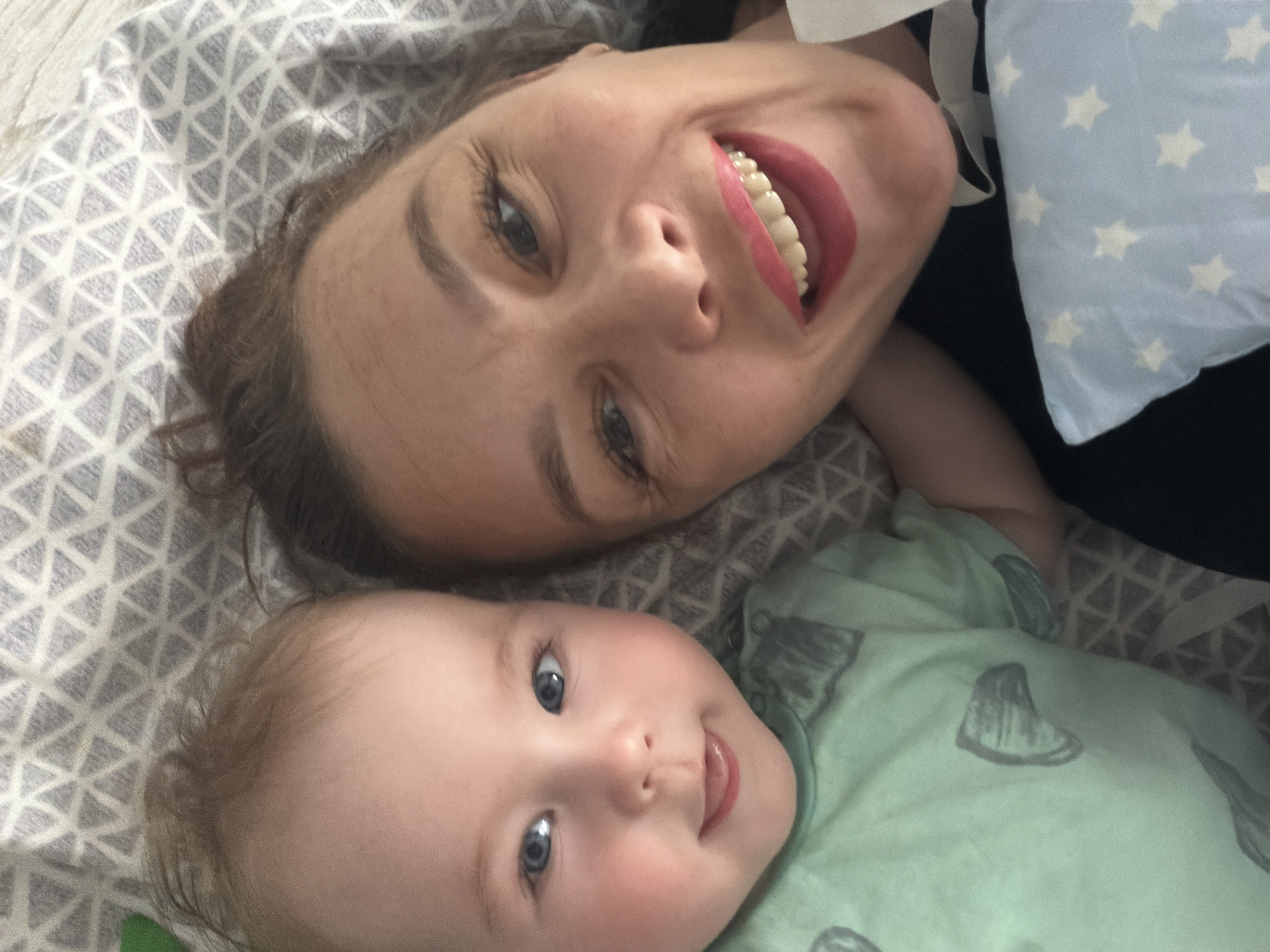 This is a close-up, overhead photograph of a woman and a baby lying down together. The woman is in the upper portion of the frame, looking up at the camera with a wide smile, her teeth visible. She has dark hair and dark eyebrows. The baby is positioned below the woman, also looking towards the camera with a slight smile and its tongue peeking out. The baby has blue eyes and light brown hair.
Both subjects are lying on a surface covered with a grey and white patterned fabric. A light blue pillow with white stars is visible to the right of the woman's head. The baby is wearing a light green onesie with a pattern of dark grey shapes, possibly representing leaves or feathers. The overall mood of the image is joyful and affectionate, with both individuals appearing happy and relaxed. The lighting suggests it might be daytime, possibly indoors. There is no visible text in the image.