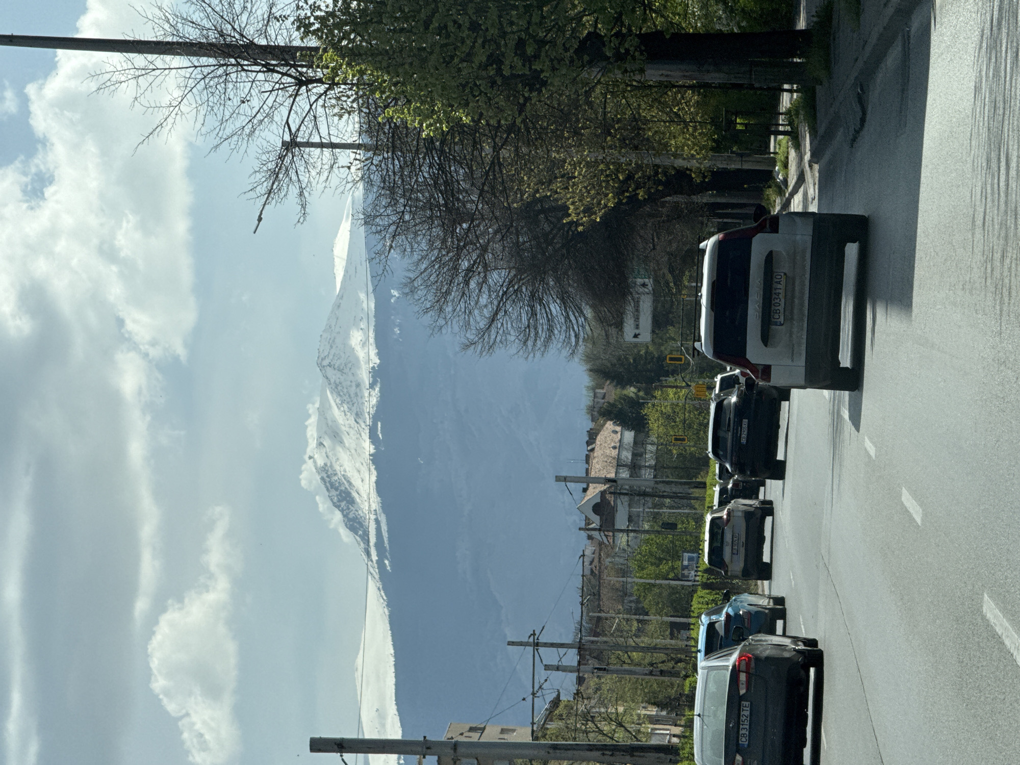The image presents an urban street scene in Sofia, Bulgaria, looking towards the majestic, snow-capped Vitosha Mountain under a partly cloudy sky. The perspective is from inside a vehicle, observing a line of cars in traffic.
In the foreground, a multi-lane asphalt road features white lane markings. Several cars are visible, including a dark grey station wagon closest to the viewer with the Bulgarian license plate "CB 3152 TE". Ahead of it, a blue car is followed by another vehicle displaying the partially visible plate "CB 2895 AE". Further along, a white Dacia SUV with the license plate "CB 0341 AO" stands out.
To the right of the road, a sidewalk is lined with trees; some are bare, indicating early spring, while others show fresh green leaves. Utility poles and overhead wires crisscross the frame. A white street sign with black text and arrows provides directions: "В. ЛЕВСКИ" (V. LEVSKI) with a right-pointing arrow and "ЦЕНТЪР" (CENTER) with a left-pointing arrow. A yellow traffic light signal is also visible. Beyond the road infrastructure, residential buildings are nestled amongst the greenery.
The background is dominated by the prominent, snow-covered peak of Vitosha Mountain, confirming the location. The sky features a mix of light blue and white clouds, indicating daylight hours, likely morning or afternoon, with clear visibility. The scene captures a typical city environment against a dramatic natural backdrop.