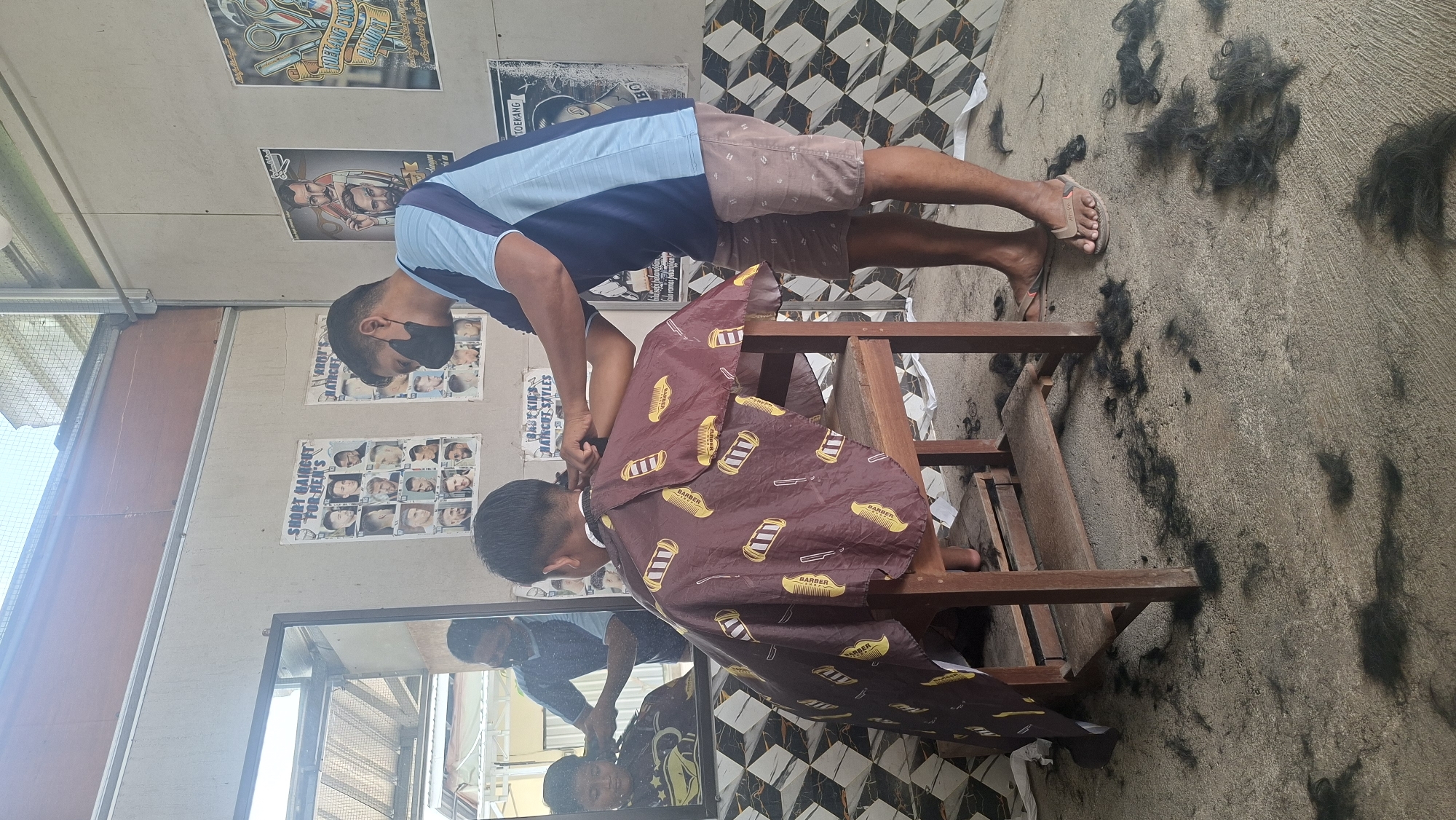 In a modest barbershop located in Bantengmati, Indonesia, a barber wearing a blue and navy striped shirt and brown shorts with a subtle pattern, and a black face mask, is cutting the hair of a young boy. The boy is seated on a wooden chair and draped with a maroon cape adorned with a repeated pattern of yellow and black combs and scissors. The barber is actively engaged in the haircut, holding clippers in his right hand and using his left hand to guide the hair.

The barbershop's walls are decorated with posters displaying various hairstyles for men and children. To the left, a large mirror reflects the scene, showing the barber from a slightly different angle and a glimpse of the street outside. The floor of the establishment is concrete and scattered with fallen hair, indicating that the haircut is in progress or has recently concluded. The overall environment suggests a functional, no-frills barbershop.