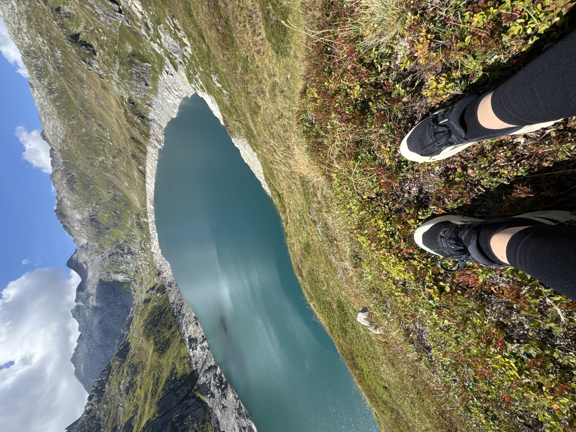 The image depicts a person's legs and feet clad in black leggings and athletic shoes, positioned on a grassy slope overlooking a large, turquoise lake. The lake is surrounded by steep, rocky mountains covered in patches of green vegetation. The sky above is blue with scattered white clouds. The foreground shows a mix of green grass and low-lying shrubs with red and yellow leaves. The perspective is from a first-person view, as if the viewer is sitting on the slope. The scene appears to be an outdoor natural landscape, possibly in the Swiss Alps near Airolo, Switzerland, given the mountainous terrain and the type of lake. There are no other individuals or structures immediately visible in the frame. The lighting suggests it is daytime, with sunlight illuminating the landscape.