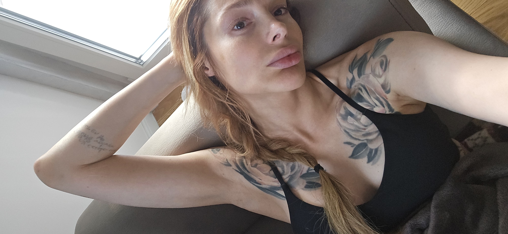 A woman lies on a grey upholstered surface, possibly a sofa or chaise, indoors. Her head is tilted toward the camera, and she rests her left arm behind her head. She has light skin and long, reddish-blonde hair styled in a loose braid that falls across her chest. She wears a black tank top. The woman has several tattoos on her upper arms and chest. Natural light filters in from a window in the background. The location cannot be definitively determined as Podgorica, Montenegro.
