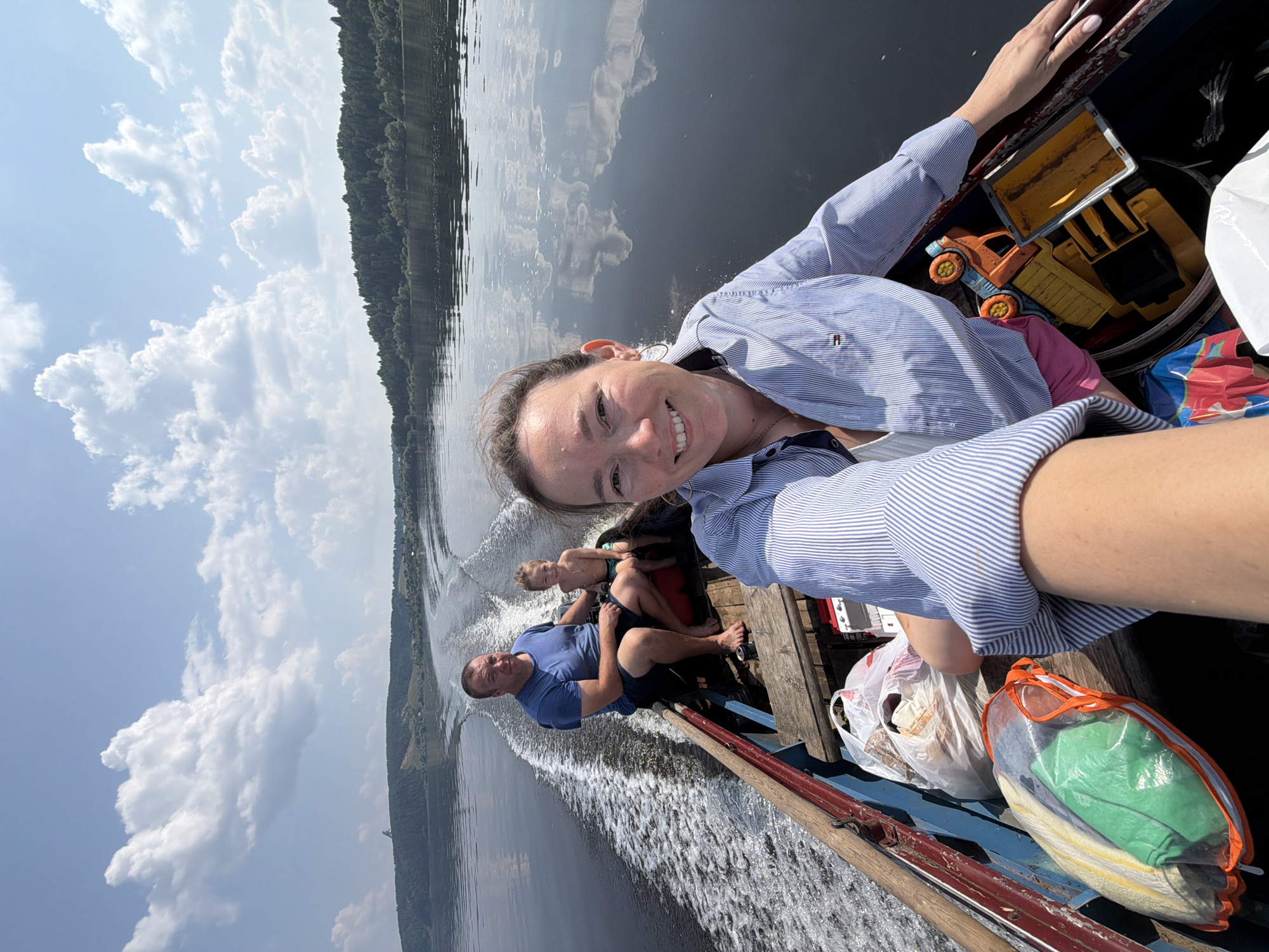 A family of three is on a boat ride on a river. The front passenger is a woman wearing a blue and white striped shirt and pink shorts, smiling at the camera. Behind her, a man in a blue t-shirt and shorts sits with a young boy wearing green shorts. The boat is moving, leaving a wake in the water. In the foreground, there are several plastic bags containing belongings, as well as a toy truck. The background shows a tree-covered hillside and a bright blue sky with fluffy white clouds. The scene appears to be in Ol'khovskoye, Russia.