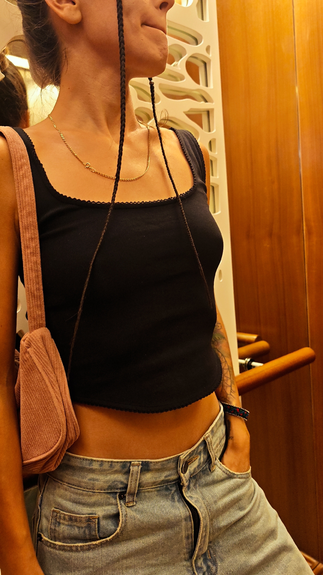 The image shows a medium shot of a person, viewed from the waist up. The person has dark hair styled in two braids that hang down in front of their shoulders. They are wearing a black tank top with a scalloped edge and light-wash denim jeans. A small, pinkish-brown purse is slung over their left shoulder. The person's right hand is tucked into the front pocket of their jeans, and a colorful bracelet is visible on their wrist. Their skin has a warm, tanned tone.
The background appears to be indoors, with a wooden wall and a decorative screen with cutout patterns. The lighting suggests it could be daytime, but the interior setting makes it difficult to determine the exact time. The overall ambiance is casual and relaxed. The image does not contain any visible text.