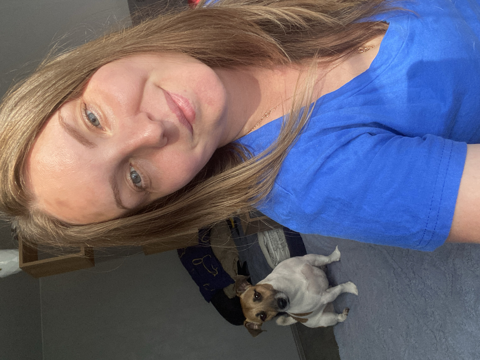 In a domestic setting, a young woman with blonde hair and blue eyes smiles at the camera, her face illuminated by natural light. She is wearing a vibrant blue t-shirt and a delicate gold necklace. To her left and slightly behind her, a small, tan and white Jack Russell Terrier sits attentively on a textured blue rug. The background features a dark gray wall and a wooden shelf with a white object, possibly a plant pot. The scene appears to be a casual selfie taken indoors. The image does not provide any specific information about the location being in the city of Noviy, Russia.