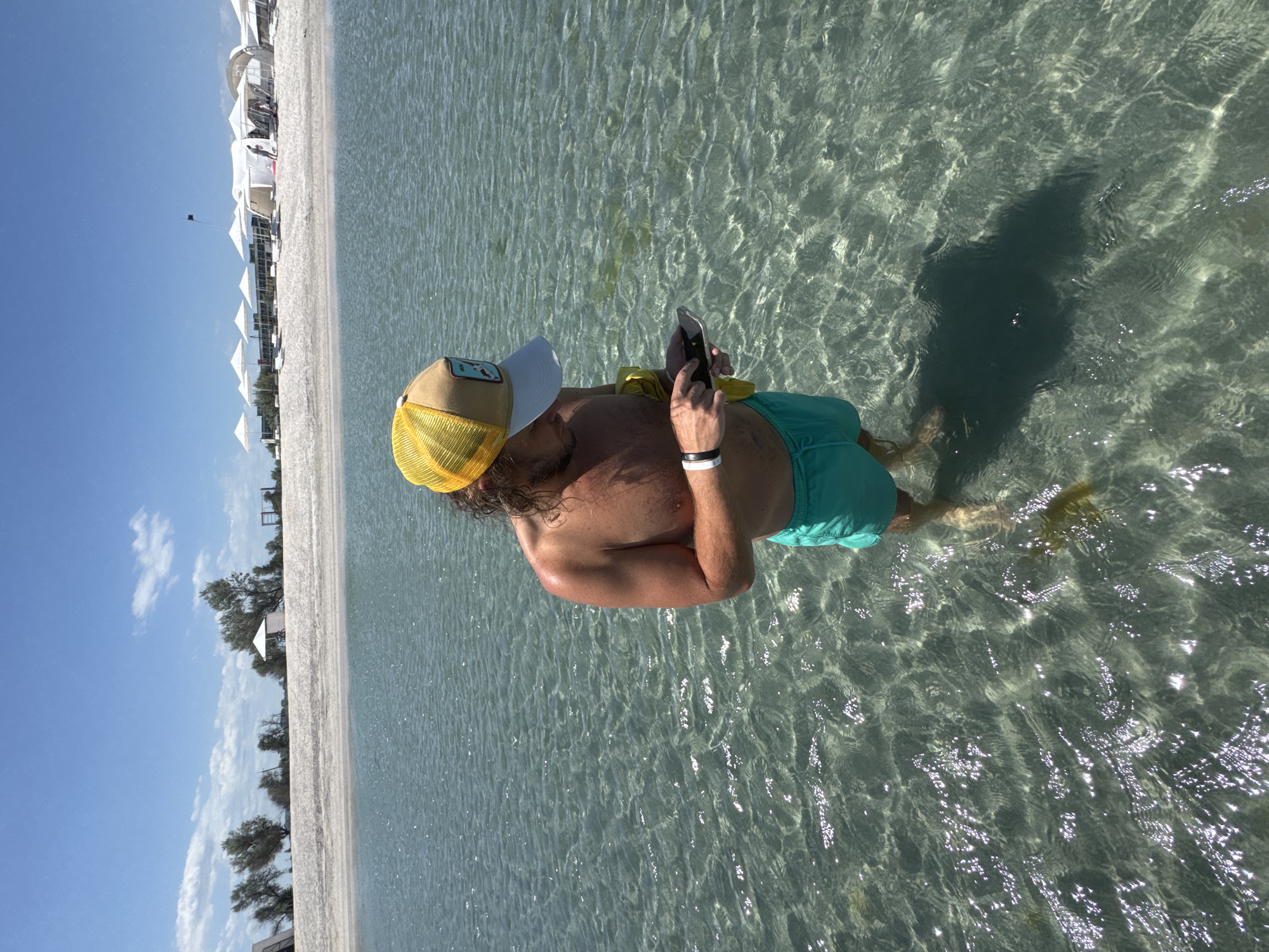 A medium, eye-level shot captures a shirtless man with a tanned complexion standing in shallow, clear water next to a sandy beach. The man wears a yellow and white baseball cap and turquoise swimming trunks. He holds a dark-colored cell phone in his hand, looking down at it with his index finger extended towards the screen. He wears a white and black band on his wrist. The sandy beach stretches along the left side of the frame. The sun reflects on the water's surface, creating a glistening effect. The location may be a beach within Мысовское сельское поселение, Ukraine.