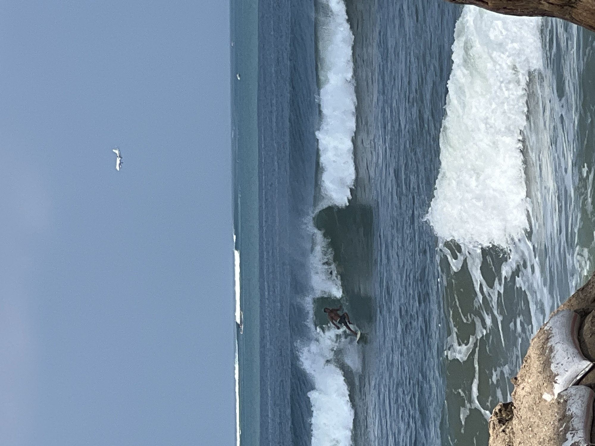 In Kuta, Indonesia, a surfer rides a breaking wave in the ocean. The surfer is a male, shirtless, wearing dark shorts, and is in a crouched position on a surfboard. The wave he is riding is a medium-sized, foamy crest. In the background, multiple rows of waves are visible, with a boat in the distance on the second row of waves. Above the ocean, a white airplane is flying in the clear blue sky. In the foreground, a rocky shoreline with sandbags is visible.