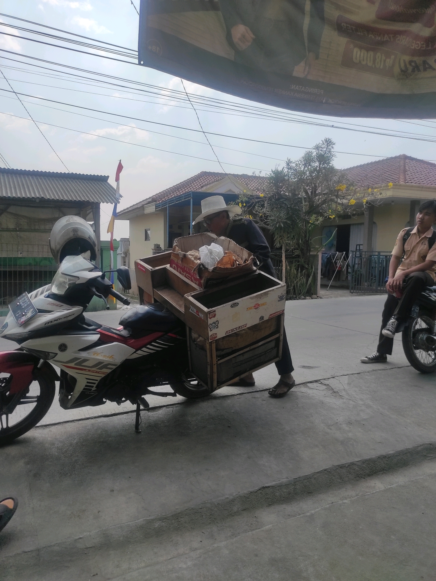 A man wearing a white hat and dark clothing is pushing a wooden cart attached to a motorcycle. The cart is laden with goods, including boxes and bags. The motorcycle, a white and red Yamaha MX-King 150, is parked on the side of a road. To the right of the scene, a young man in a school uniform is sitting on another motorcycle. The background shows houses with tiled roofs and trees. The sky is overcast. The scene appears to be a street in Ngamprah, Indonesia, likely in a residential area given the houses and road conditions. The overall impression is of a street vendor or delivery person at work.