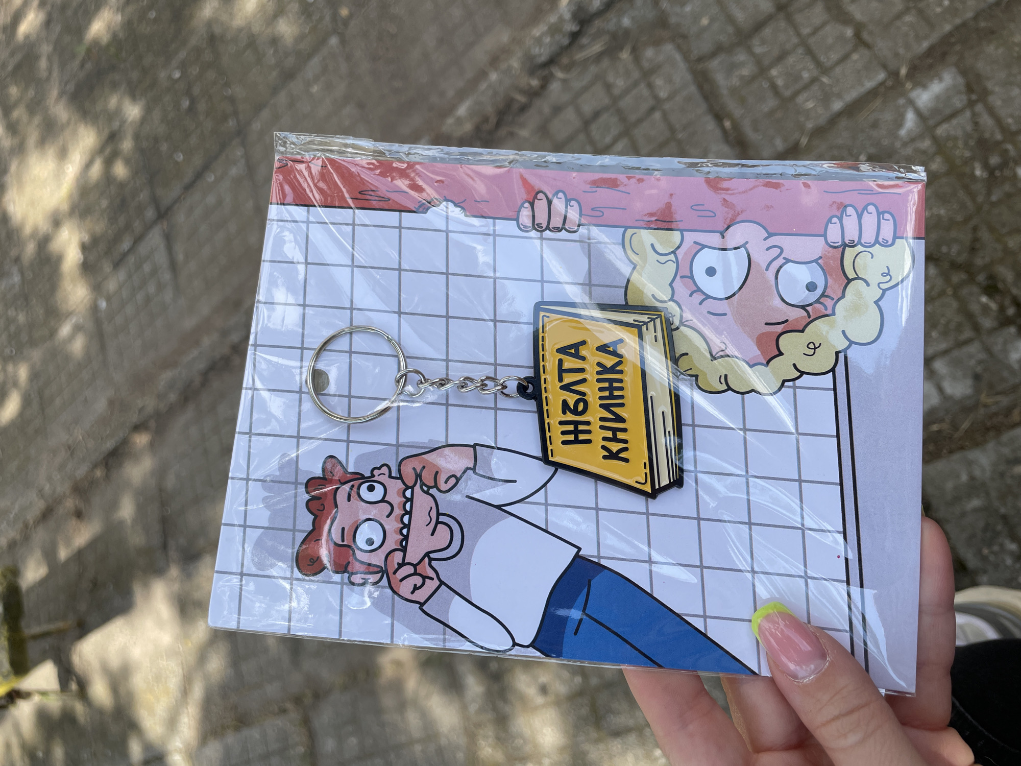 A keychain with a yellow book charm featuring Cyrillic text is attached to a card depicting two cartoon characters. The card has a white grid background and a red border at the top. One character is a man with red hair, wide eyes, and his fingers stretched to his cheeks, making a distorted face. He is wearing a white shirt and blue pants. Above him, peeking over the red border, is a blond-haired character with bulging eyes and a surprised or distressed expression. The keychain and card are enclosed in clear plastic packaging. The scene appears to be outdoors, with a blurred background of a paved surface that resembles cobblestones or a sidewalk. The lighting suggests it is daytime, likely sunny, with dappled shadows. The location is Dimitrovgrad, Bulgaria. The text on the yellow book charm reads "ЖЪЛТА КНИЖКА" which translates to "Yellow Book".