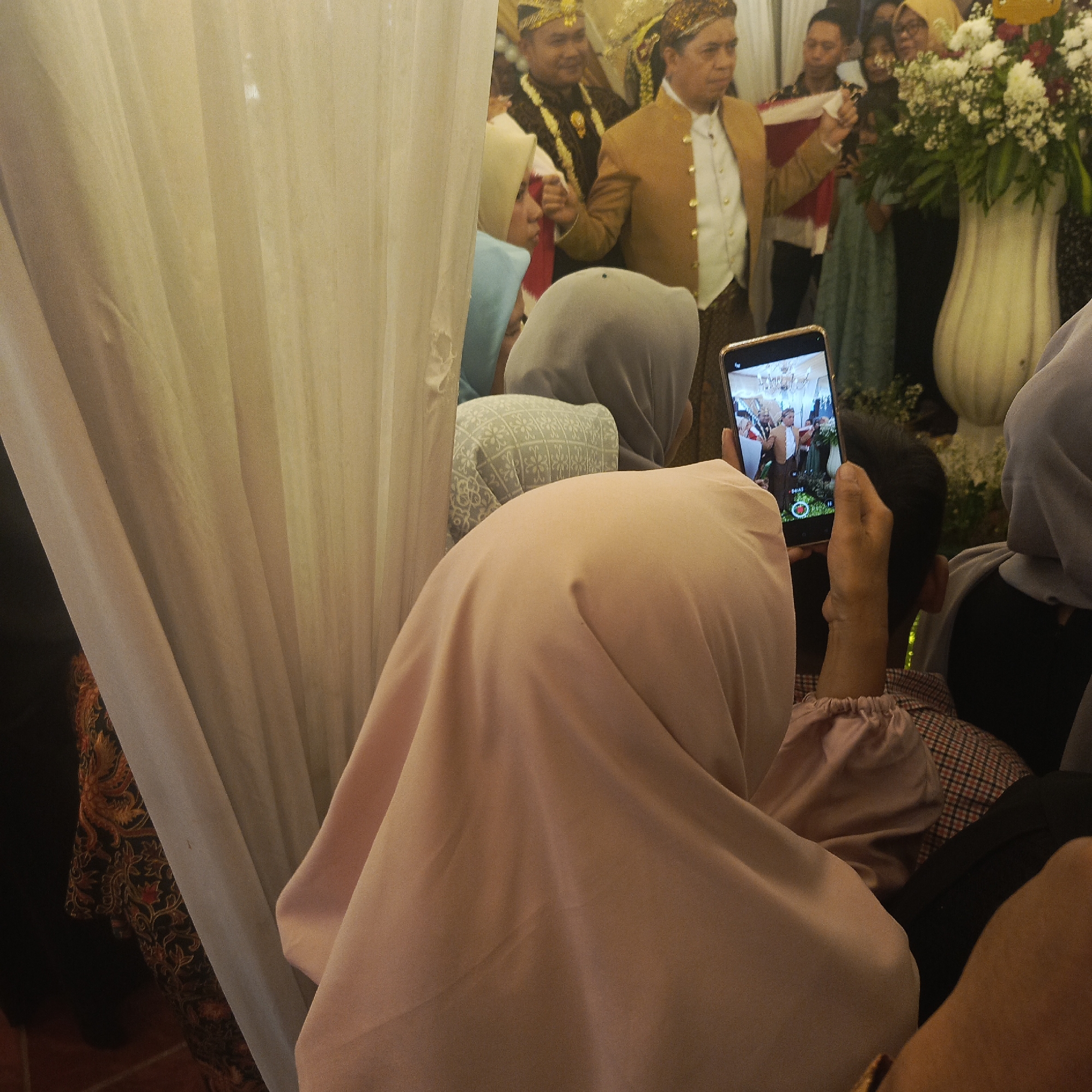In South Jakarta, Indonesia, a wedding ceremony is underway, with guests gathered around. A person in the foreground is holding a smartphone, capturing the event. The phone displays a scene of two men in traditional attire, presumably the bridegroom and an elder, standing next to a large floral arrangement. The man on the left wears a dark, ornate jacket and a gold headdress, while the man on the right is dressed in a gold jacket and a traditional cap. Behind them, other guests are visible, some wearing headscarves. The environment appears to be indoors, with soft lighting and draped fabric suggesting a decorated venue. The overall atmosphere is one of celebration and family gathering.