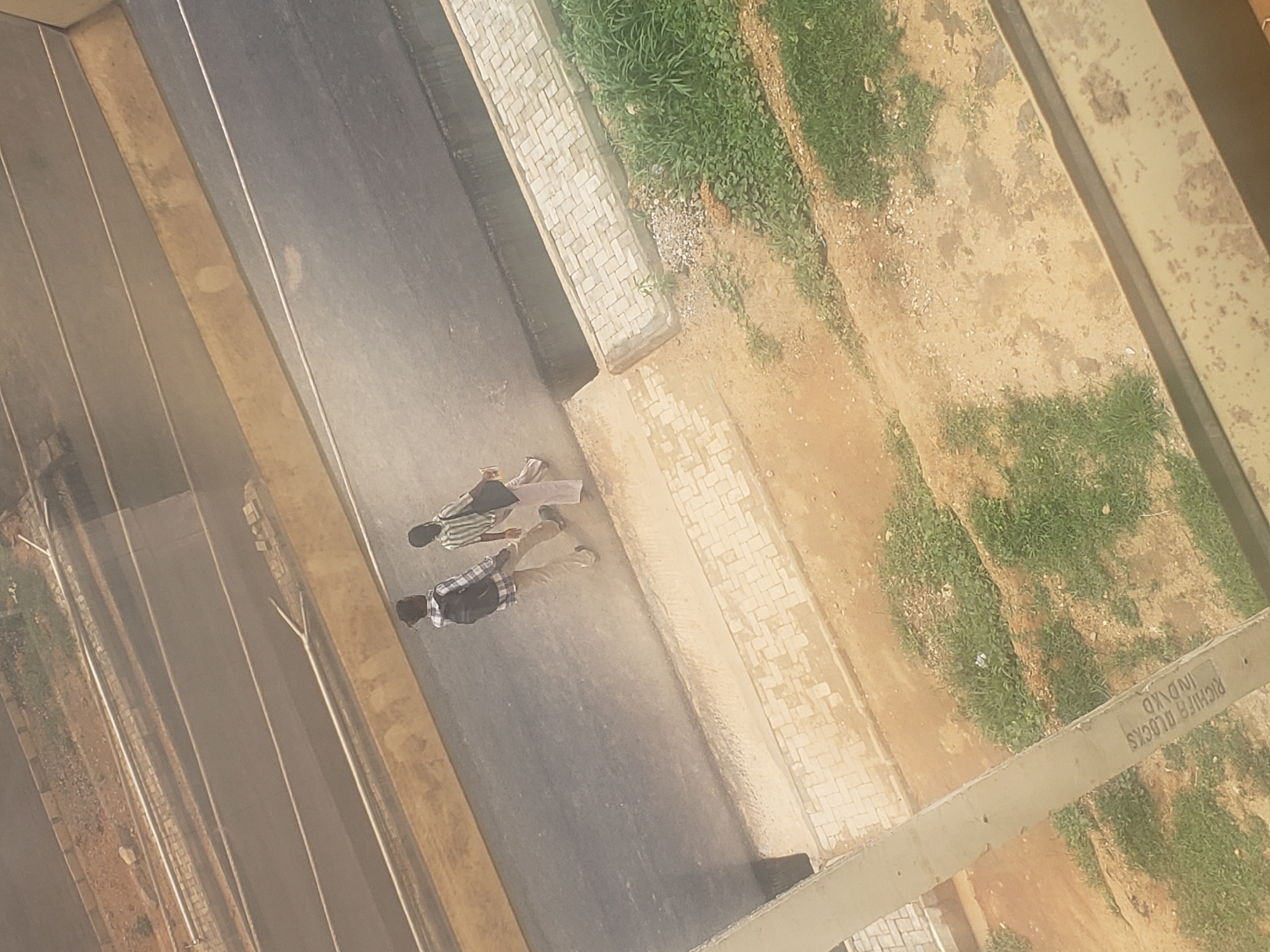 Two individuals, one wearing a plaid shirt and the other a striped shirt, walk across a paved road. Both appear to be carrying bags. The road is bordered by a yellow barrier on one side and a sidewalk with interlocking pavers on the other, which transitions into a grassy area. The scene appears to be an outdoor urban environment in Gumel, Nigeria. The perspective is from an elevated position, looking down at the road and surroundings.