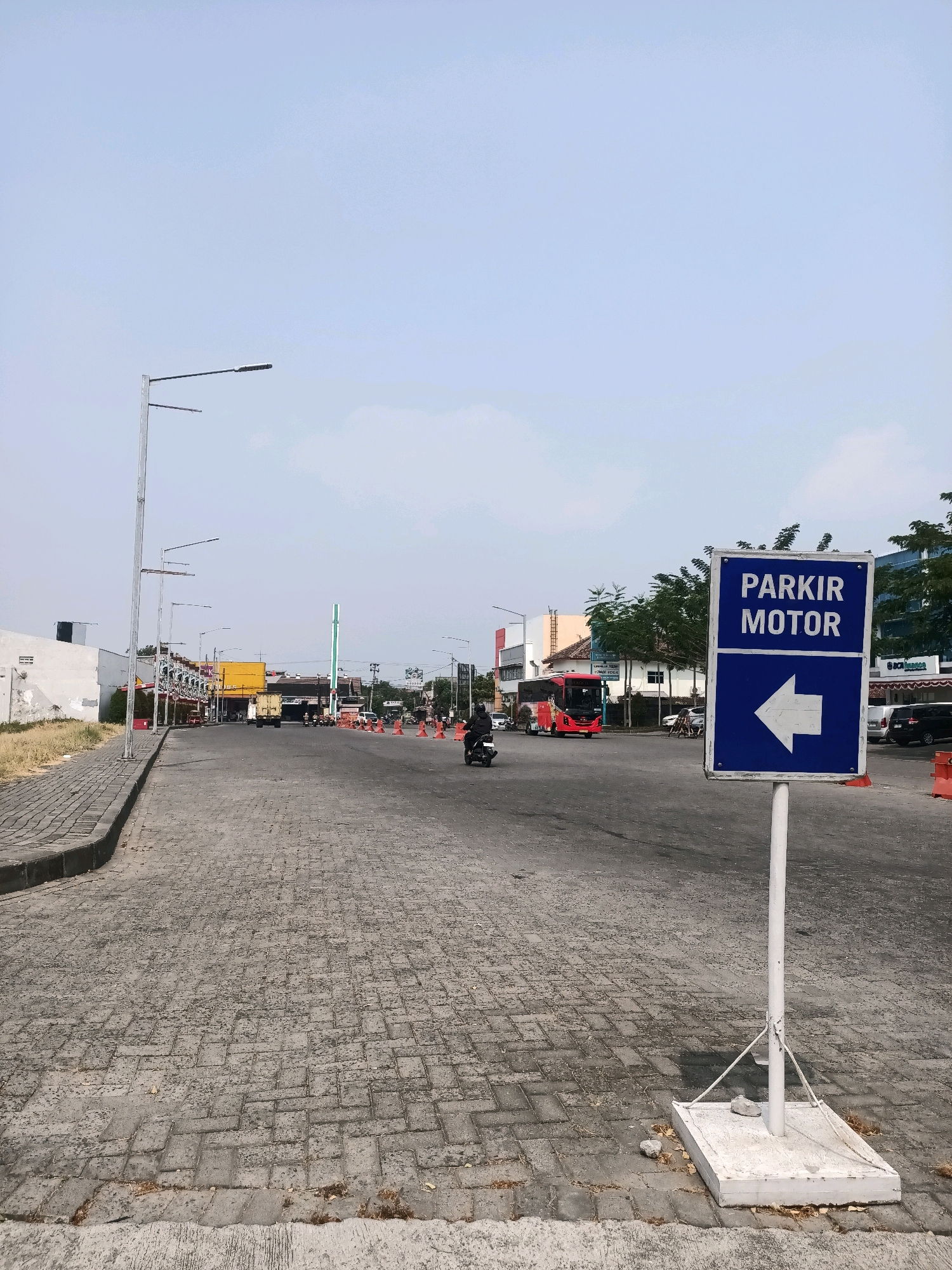 In the city of Grogol, Indonesia, a street scene unfolds under a clear sky. The foreground is dominated by a textured paved surface, appearing to be interlocking concrete blocks. To the right, a white pole supports a blue and white directional sign indicating "PARKIR MOTOR" (Motorcycle Parking) with a white arrow pointing left. The street itself is relatively wide, with a curb on the left side that transitions into a grassy verge.
Further down the street, traffic is visible. A person on a motorcycle rides away from the camera. In the mid-ground, a red bus is traveling in the opposite direction. Various other vehicles, including cars and trucks, are present on the road. Several orange traffic cones are strategically placed along the right side of the road, suggesting ongoing work or a designated area. Buildings line the sides of the street, appearing to be commercial establishments, some with awnings. Streetlights are visible along the road, and trees with green foliage are scattered throughout the scene, particularly in the background. The overall environment suggests a developed urban area with ongoing activity.
