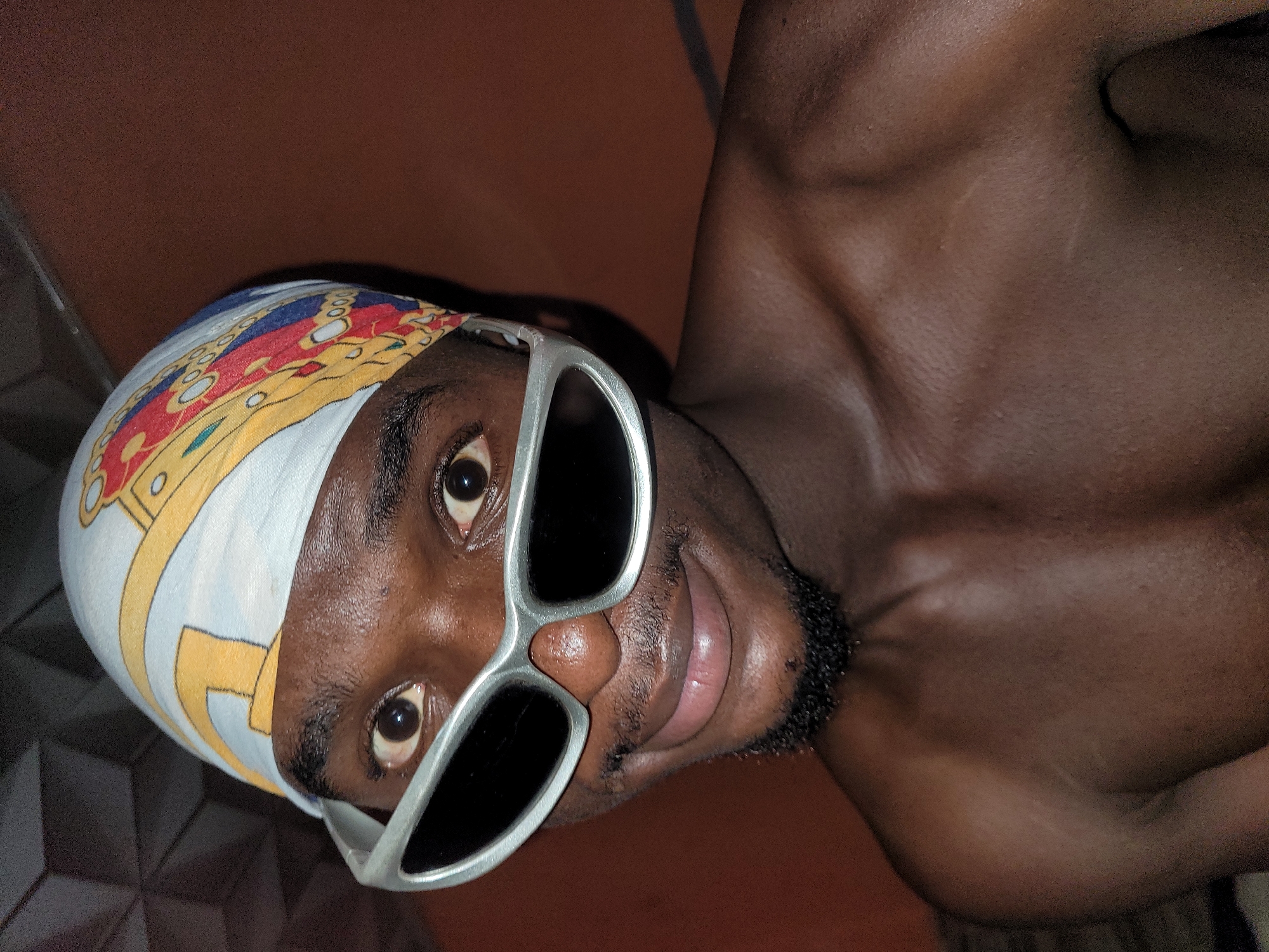 A close-up, eye-level, medium shot captures a young Black person wearing a bandana with a gold and red pattern and silver, wraparound sunglasses. The person's head is tilted slightly to the right, and they are looking directly into the camera with a subtle smile. Their chest and shoulders are visible, with no shirt. The background consists of a patterned wall and a brown, textured wall. The image appears to be taken indoors. The subject is located in Bukuru, Nigeria.