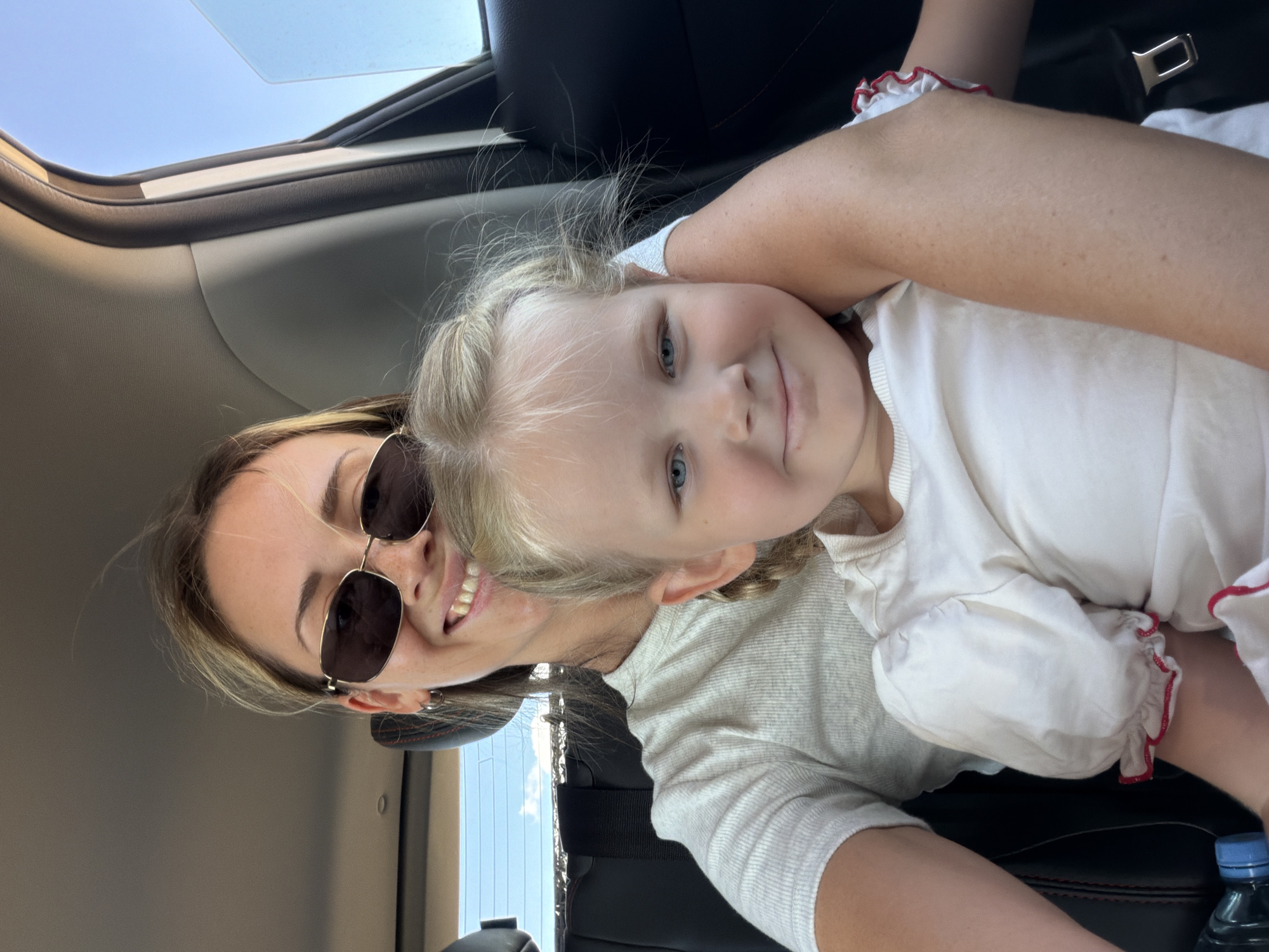 In the city of Ternovka, Russia, a woman and a young girl are seated in the back of a vehicle, likely for a selfie. The woman, positioned behind and slightly to the left of the girl, wears sunglasses and a white t-shirt. She is smiling broadly, with her hair partially obscuring her face. The girl, positioned in the foreground, looks directly at the camera with a gentle smile, her blonde hair styled in braids. She is wearing a white shirt with red trim on the sleeves. The interior of the car is visible, including dark upholstery and a seatbelt. The window of the car is open, revealing a clear blue sky with some distant clouds. The lighting suggests it is daytime.