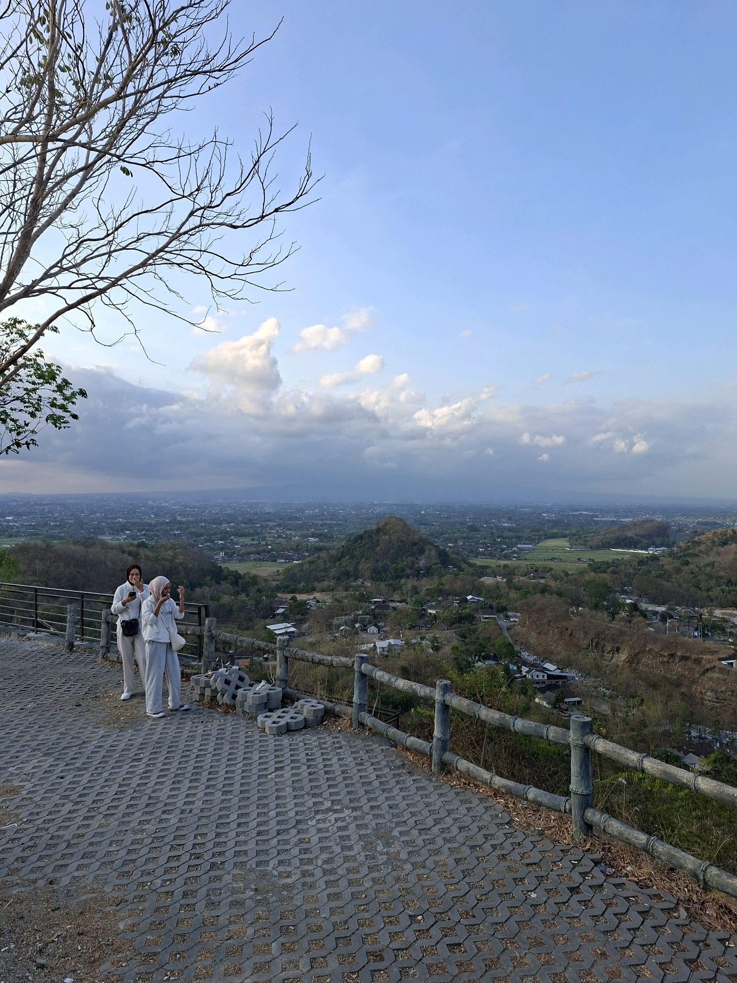 The image captures a scenic overlook in Pleret, Indonesia, with a panoramic view of a verdant landscape under a partly cloudy sky. In the foreground, two women stand on a paved walkway made of interlocking grey blocks. The woman on the left is wearing a light-colored top and pants, holding her phone and looking towards the landscape. The woman on the right, wearing a hijab and a matching outfit, is also holding her phone and appears to be taking a photo or video. They are positioned near a rustic wooden railing that lines the edge of the overlook. A pile of grey interlocking concrete blocks is stacked to their right.
The middle ground reveals a sprawling town with houses nestled among rolling hills and lush greenery. A distinctive conical hill rises from the landscape in the distance. The background is dominated by the expansive sky, with a mix of white, puffy clouds and areas of clearer blue, suggesting either early morning or late afternoon. The overall atmosphere is serene and picturesque, offering a wide vista of the Indonesian countryside.