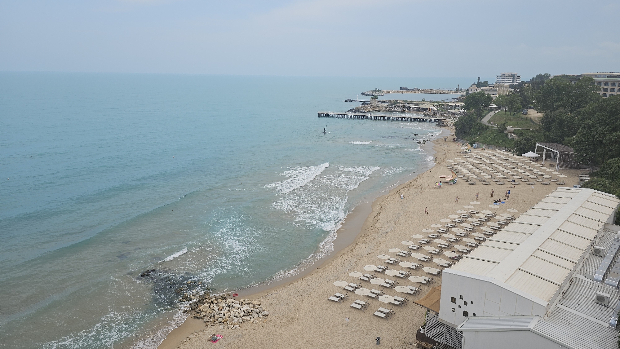 An aerial long shot captures a vibrant beach scene on an overcast day in Varna, Bulgaria. The turquoise-blue sea, calm with gentle waves, stretches into the horizon under a hazy sky. A sandy beach, dotted with numerous rows of neatly arranged white sun loungers and umbrellas (some open, some closed), borders the coastline.
Several people are present on the beach and in the water. Individuals are seen walking along the shore, a small group appears to be playing a game in the sand, and others are relaxing near the loungers. In the sea, a lone person is paddleboarding, and two small buoys are visible further out. Rocky sections are interspersed along the shoreline and in the shallow waters.
Towards the right foreground, a large, multi-level building with a white roof and visible air conditioning units stands adjacent to the beach. Further along the coastline, a long wooden pier extends into the sea, with more substantial stone and concrete jetties and structures visible in the distance. The background reveals a lush green hillside densely covered with trees and bushes, featuring paths and additional buildings, likely hotels or resorts, overlooking the beach. The overall atmosphere is one of serene leisure and recreation at a coastal resort. No discernible text is visible.