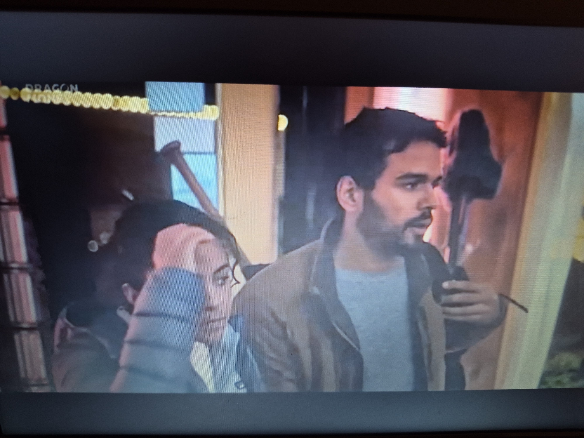 The image shows a close-up of a television screen displaying a scene with two people. The person on the left is a woman with dark hair, wearing a blue jacket. The person on the right is a man with dark hair and a beard, wearing a grey t-shirt and a brown jacket. He is holding an object in his right hand. The background appears to be indoors with some artificial lighting, suggesting it could be a set or a studio. The top left corner of the screen shows a logo that reads "DRAGON" in yellow lettering, followed by a series of yellow dots. There is no visible indication of a specific location within the city of Sandwell, United Kingdom.