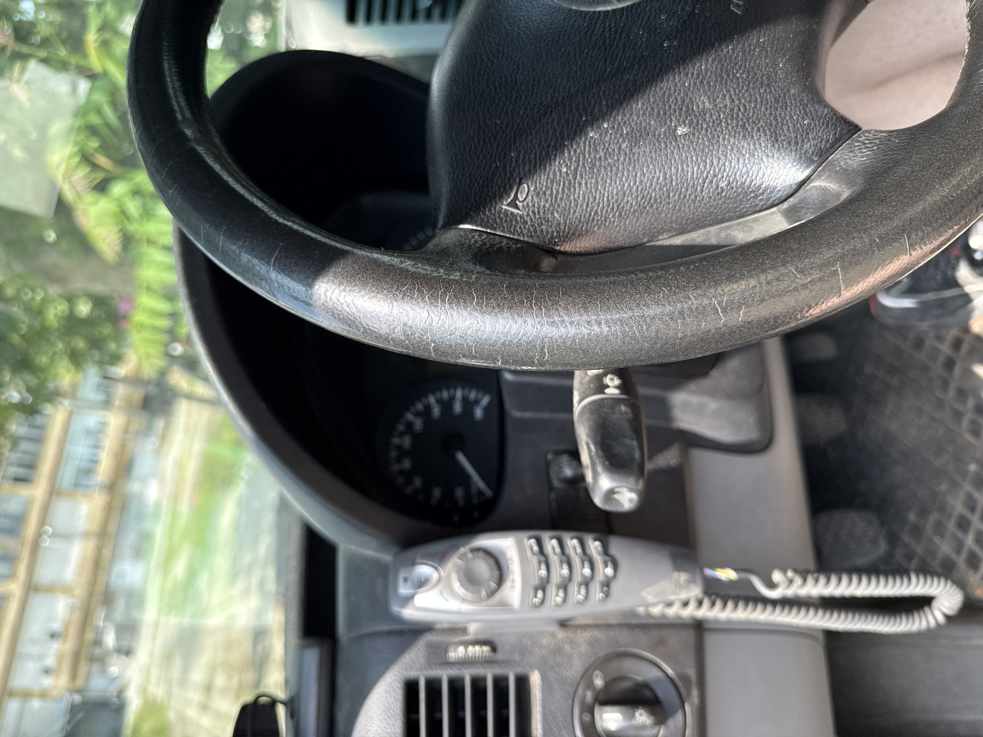 The image captures the interior of a vehicle, predominantly focusing on the dashboard and steering wheel from the driver's viewpoint. The steering wheel is made of a dark material, possibly leather or a synthetic equivalent, and exhibits considerable wear and cracking on its rim. A small symbol resembling a post horn, commonly used for a horn button, is visible on the steering wheel's hub.
Below the steering wheel, part of the instrument cluster is visible, including a speedometer with its needle resting at zero, indicating the vehicle is stationary. To the right of the cluster, a control stalk, likely for turn signals or wipers, is present.
A prominent feature on the dashboard is an older model car phone or handset. It has a central rotary selector button and a numeric keypad with visible numbers (1-9, *, 0, #). A distinctive white coiled cord connects the handset. Further down the dashboard, an air vent with horizontal slats and a circular control knob are discernible. The driver's floor mat shows a dark, textured pattern.
Through the windshield, a bright, blurred outdoor scene is visible. It shows what appears to be buildings with windows and a green area, possibly grass or trees, under direct sunlight, suggesting it is daytime. The strong sunlight creates clear shadows within the vehicle's interior.
The vehicle appears to be an older model, possibly a utility van or car, given the style of the dashboard and the integrated car phone. The location context for this media file is Stara Zagora, Bulgaria.