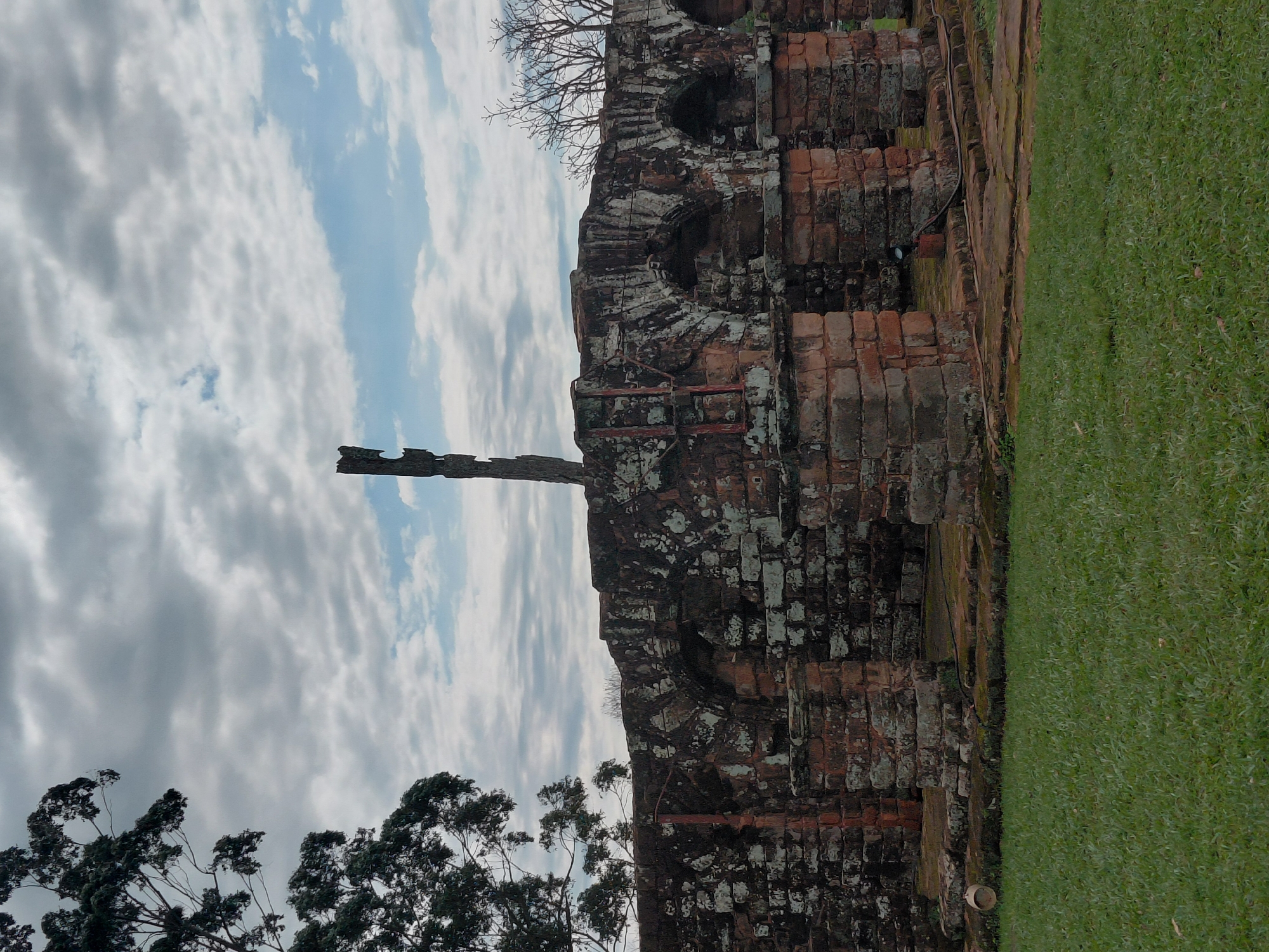 The image depicts the ruins of the Jesuit Mission of La Santísima Trinidad de Paraná, located in Trinidad, Paraguay. The foreground is a well-maintained grassy lawn. Behind the lawn, the weathered stone ruins of a historical structure are visible. The ruins feature brickwork and arched openings, some of which are partially obscured by moss and discoloration, indicating their age. A tall, weathered wooden post stands prominently behind the central section of the ruins. The background is dominated by a cloudy sky with patches of blue visible. On the left side of the image, a portion of a large tree with dark foliage is present. The overall scene conveys a sense of historical significance and natural decay.