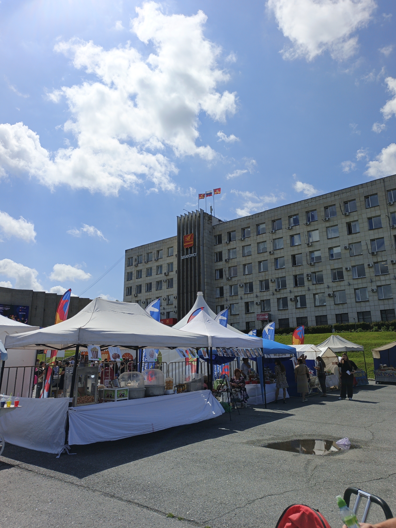 The image depicts an outdoor market scene in front of a large, multi-story building with a prominent vertical structure. The building appears to be a municipal building, as evidenced by the inscription "ЗЛАТОУСТ" (Zlatoust) and its architectural style. Two flags are visible atop the vertical structure, and several flags on poles are also present around the market stalls.
The market itself consists of several white tents and awnings, suggesting a temporary event. Inside the tents, various goods are displayed, and vendors appear to be interacting with customers. One stall prominently features equipment for making popcorn and cotton candy, with colorful bags of snacks also visible. People are seen browsing the stalls and walking around the market area.
The environment is an outdoor paved area, with some grass visible on a slope behind the market. The sky is blue with scattered white clouds, indicating fair weather. A small puddle of water is present on the pavement in the foreground, reflecting the sky and surrounding elements. The overall scene suggests a lively community event taking place on a sunny day in the city of Zlatoust, Russia.