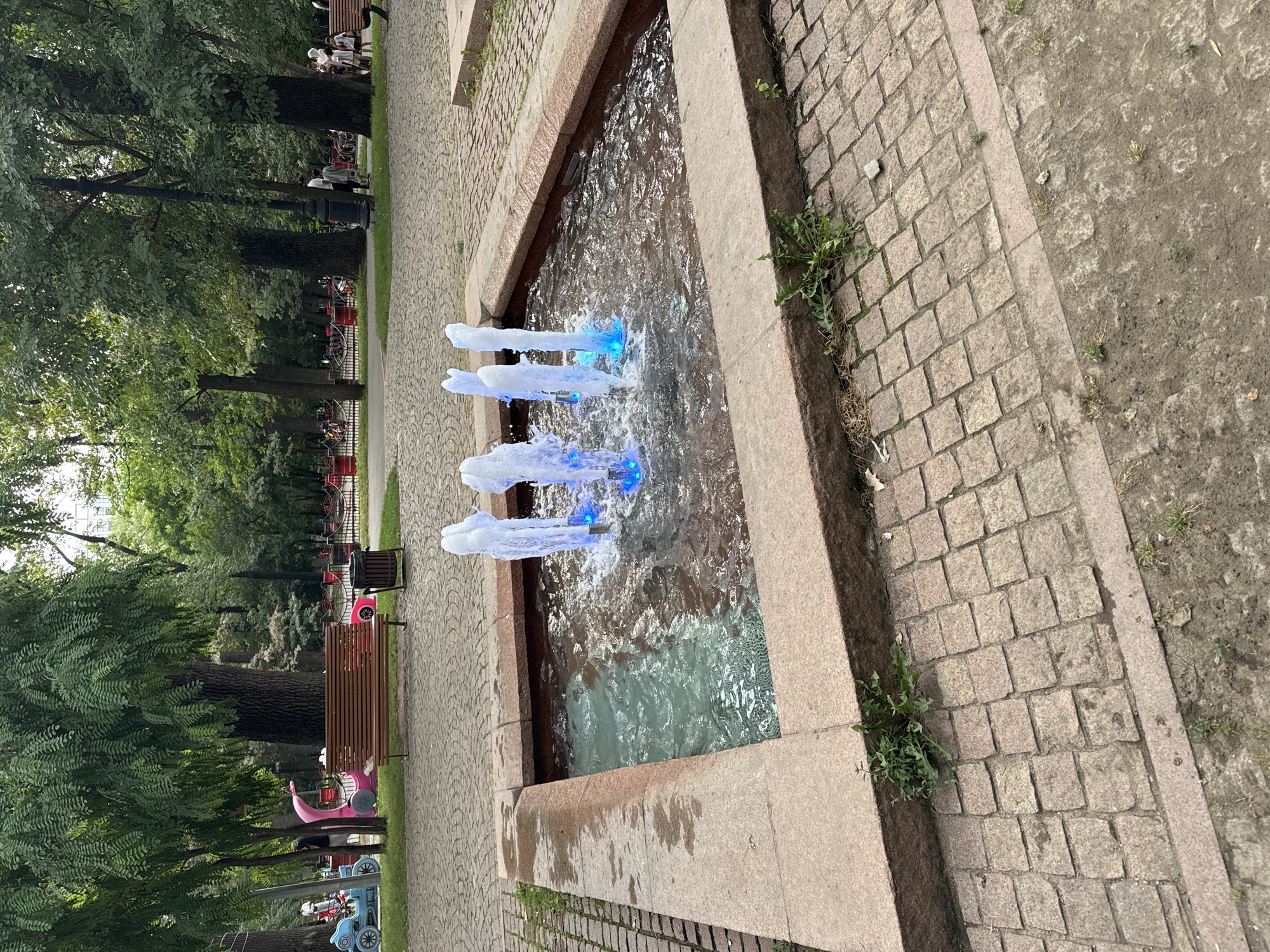 The image depicts a public park area with a rectangular water fountain in the foreground. The fountain has multiple jets of water illuminated by blue lights. The fountain is surrounded by a paved walkway made of cobblestones and granite. In the background, there are mature trees, park benches, a trash can, and several people enjoying the park. To the left, there are some children's rides, including a blue vehicle and a pink carriage. Further in the background, there is a fence with red banners and more trees. The scene suggests a pleasant, casual atmosphere in a well-maintained park.