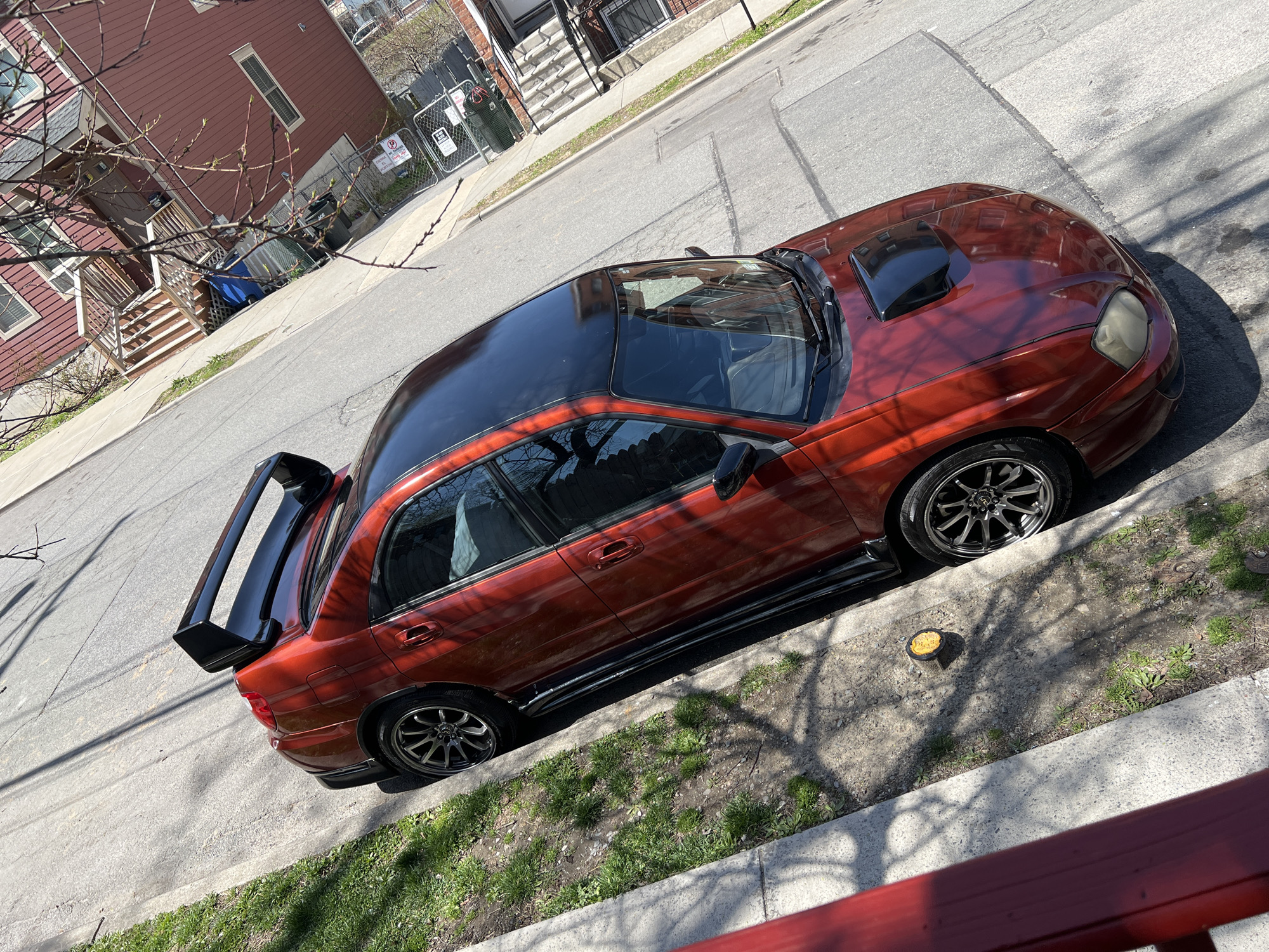 The image shows a modified, red Subaru Impreza WRX sedan parked on the street in a residential area of the City of Newburgh, New York. The car has a black roof, hood scoop, side skirts, and rear spoiler. It's parked alongside a curb with a strip of grass and sidewalk. To the left, there are red and brown multi-story buildings, trees, and a paved road. The setting is a sunny day. Near the front passenger side tire, there is an open container with a yellow food item inside. The angle of the photo is from above and to the side, possibly taken from a higher floor of a building.