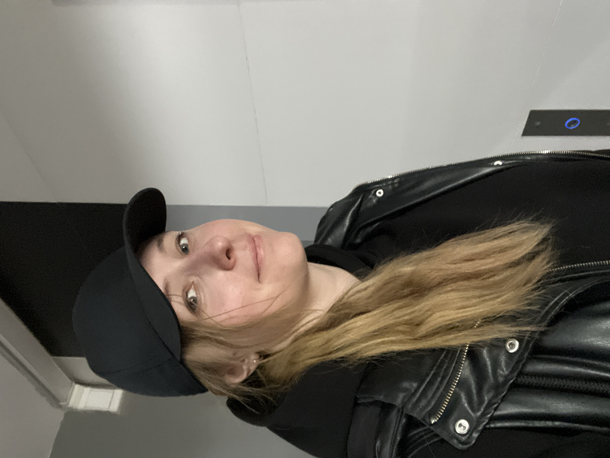 An individual is captured in an indoor setting, likely in Mytishchi, Russia. The person, appearing to be an adult, is looking directly at the camera with a neutral to slightly smirking expression. They have long, light brown or blonde hair styled over their shoulders.
They are dressed in casual attire, featuring a black baseball cap, a black hooded sweatshirt, and a black leather jacket adorned with silver zippers and snaps. The background consists of a plain light grey wall, with a darker grey or black element visible along the bottom edge. On the wall behind and to the right of the person, there is a distinct black rectangular panel that features a circular blue illuminated light and a small black dot, strongly suggesting an elevator call button or a similar control system within a building. The lighting is consistent with an indoor environment. No visible text is present in the image.
