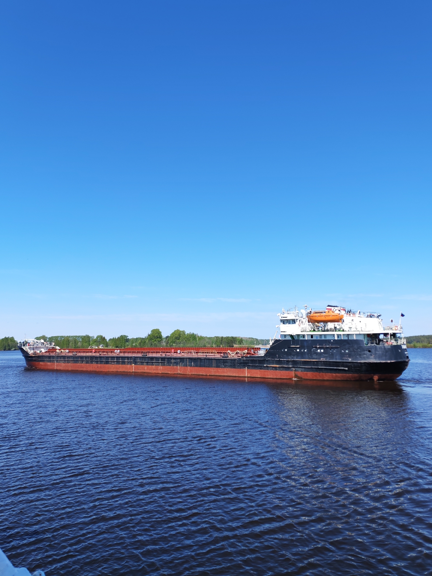 A large cargo ship navigates a wide river under a clear, cloudless blue sky. The ship is long and low-slung, with a red waterline and a dark upper hull. The ship appears to be sailing in the city of Белоусово, Russia.
