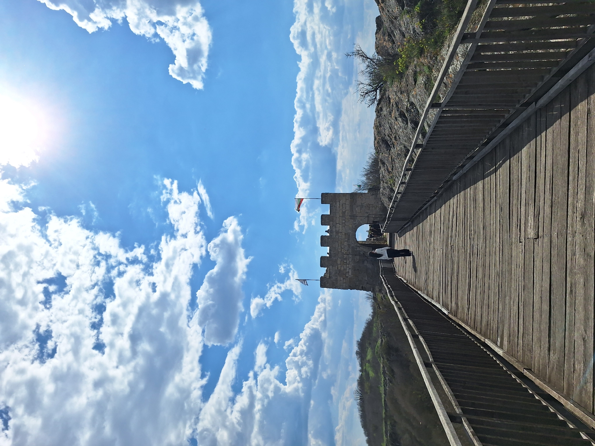 The image presents a view along a wooden bridge leading to a stone structure, likely part of the Ovech Fortress in Provadia, Bulgaria. The bridge has wooden planks and railings on both sides. A person is walking across the bridge towards the structure. The stone structure appears to be an entryway with an arch. Two flags are atop the fortress. The sky occupies a significant portion of the image, filled with puffy white clouds against a clear blue backdrop. On one side of the bridge, there is a rocky cliff face, while the other side reveals a green, forested area. The sun is visible in the upper left corner, casting a bright light.
