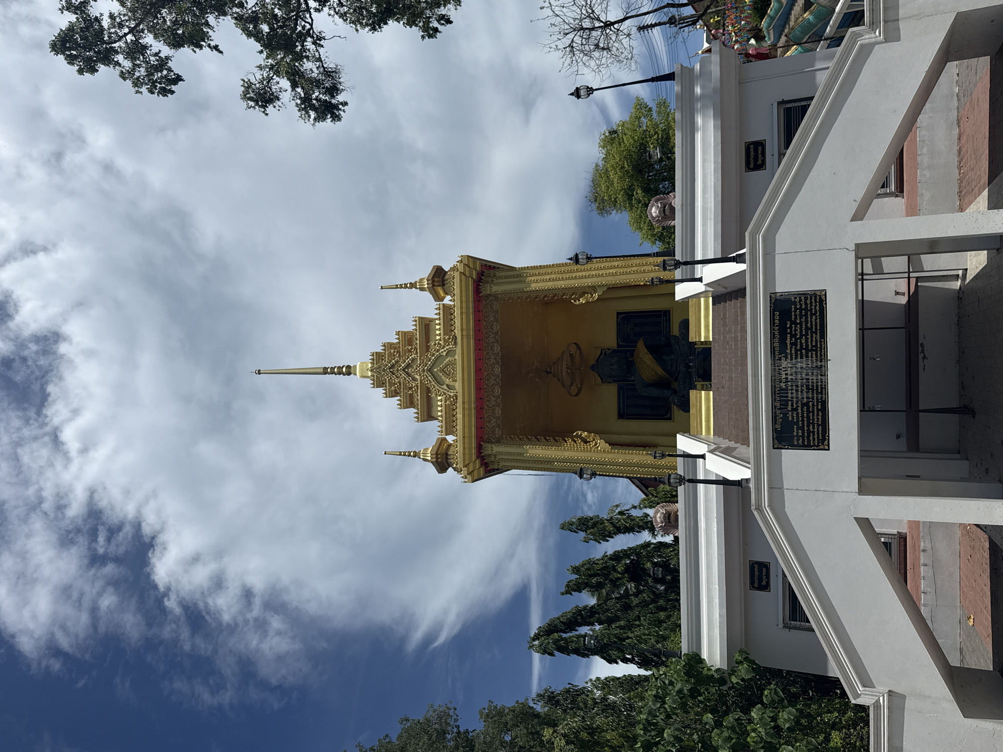 This is a low-angle, full-shot photograph of a golden temple structure in Baan Mae Nam, Thailand. The temple is ornate, with tiered roofs and intricate gold detailing. A large, dark statue of what appears to be a Buddha is seated within an alcove at the front of the temple. The temple is situated on a raised platform, accessible by a white staircase with red brick accents. There are several tall, green trees surrounding the temple, and a bright blue sky with white clouds is visible above. A black plaque with Thai script is mounted on the white wall below the staircase. Two black lampposts are positioned on either side of the stairs leading up to the temple. To the right of the main temple structure, part of a colorful staircase with blue and green accents can be seen.