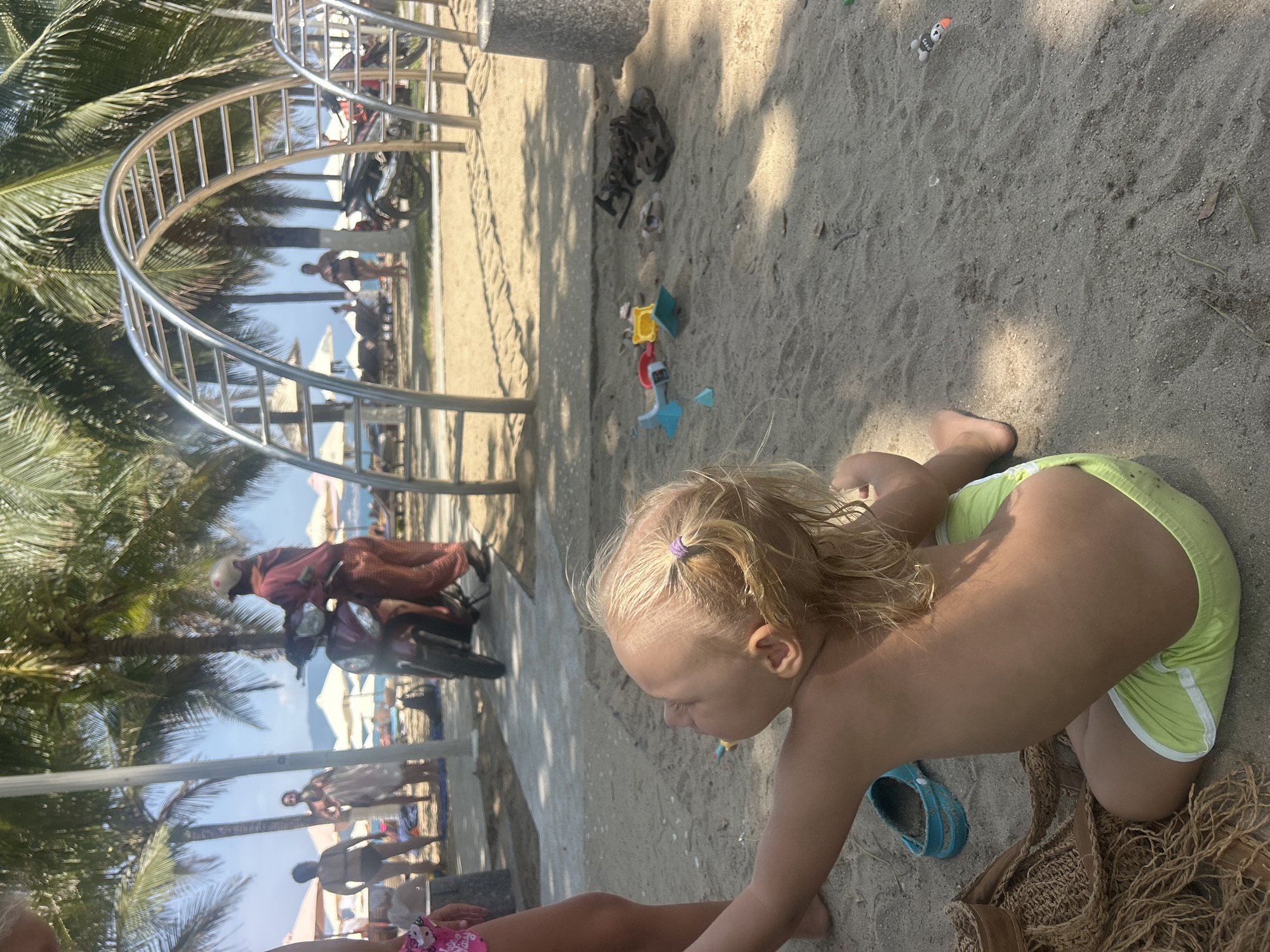 A toddler kneels on a sandy beach in Nha Trang Ward, Vietnam. The child wears light green shorts with white trim and a purple hair tie, and is looking down with their hand extended. To the child's right is a blue sandal and a woven bag. A woman's leg and foot, wearing a pink swimsuit bottom, are visible in the lower left corner. In the background, there are children's plastic toys scattered on the sand. A metal archway stands on the left side of the image, with palm trees behind it. Beyond the trees and archway, figures and white parasols are visible on the beach, with the ocean in the distance. To the left, a person on a motorcycle is visible.