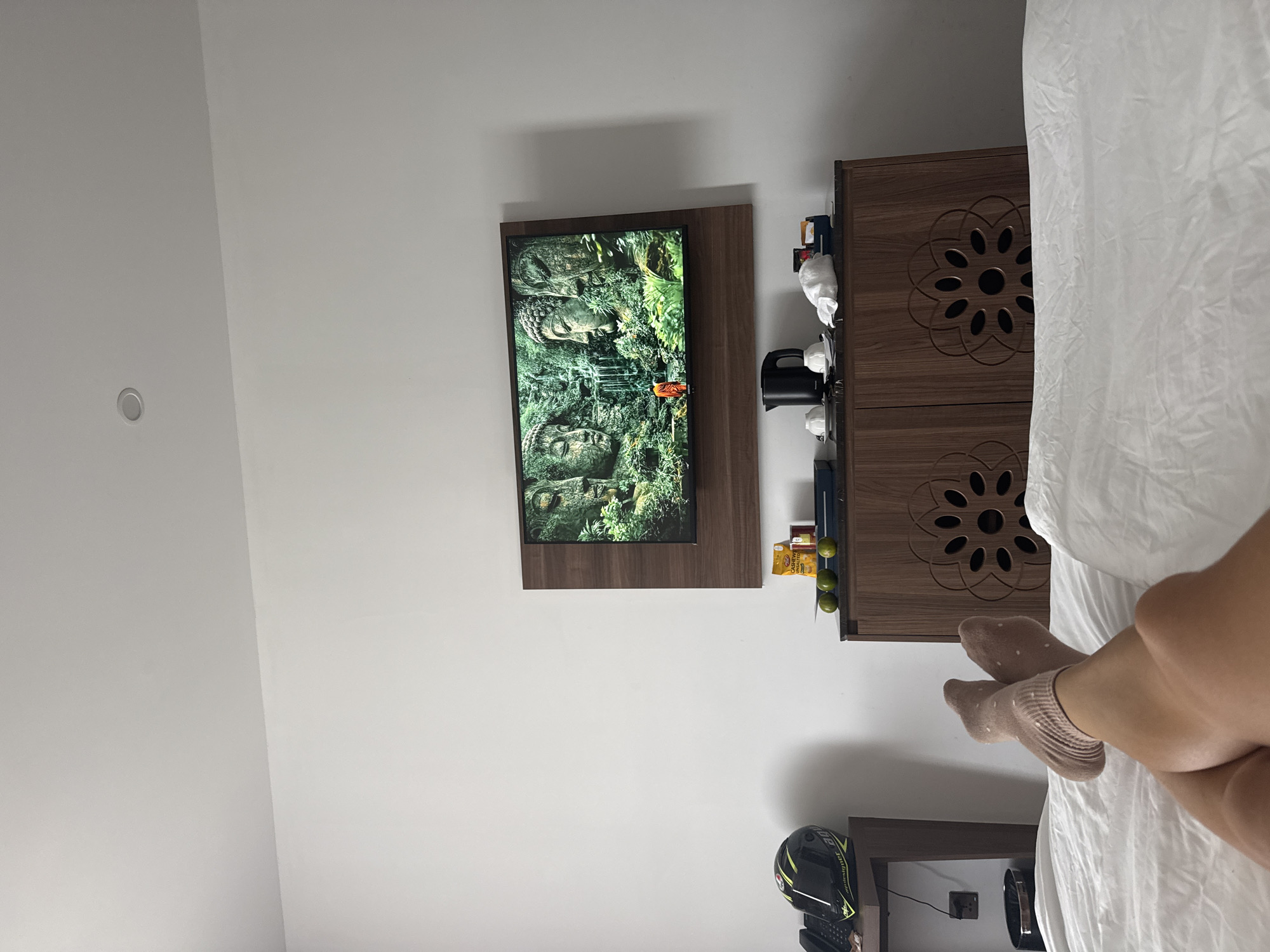 A bedroom scene in a location possibly in Đà Nẵng, Vietnam, shows a wall-mounted television displaying a video or image with a buddha-like figure. To the right is a dark wood cabinet with carved floral patterns. A black electric kettle and other items sit on a shelf above the cabinet. Below, there is a black motorcycle helmet with yellow accents on a small table. A person lies on a bed with a white sheet, legs extending into the frame. The person wears pale pink socks with white polka dots.