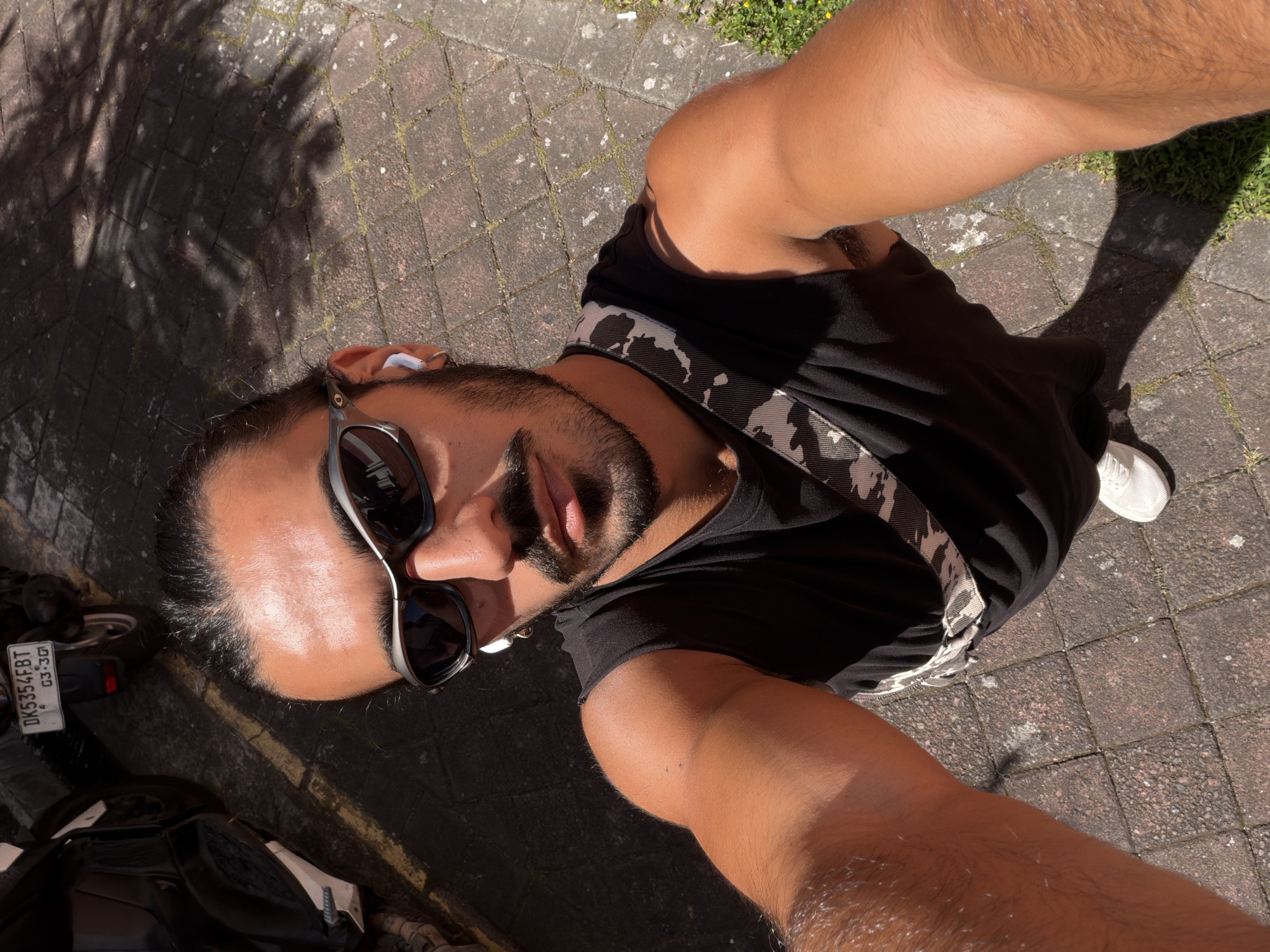 A selfie shows a person with dark hair pulled back and sunglasses on. They are wearing a black sleeveless shirt and a crossbody bag with a camouflage pattern. White earbuds are visible in their ears. The person's right arm is extended toward the camera. The background consists of a paved walkway with shadows from foliage overhead, and two motorcycles parked on the left side. A license plate on one motorcycle reads "DK5354FBT 03-30". The scene appears to be in an outdoor urban setting, possibly North Kuta, Indonesia, given the license plate format.