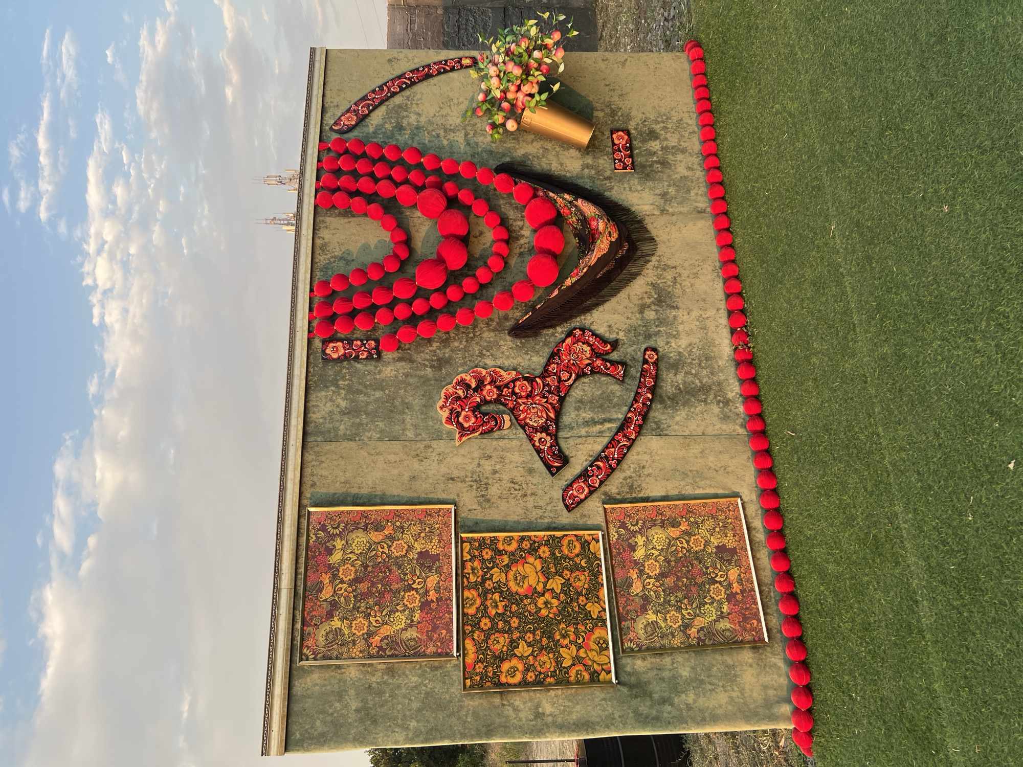 A decorative outdoor display features a large, green, fabric-covered wall adorned with colorful, folk-art-inspired elements. Three framed square pictures with intricate floral and bird patterns hang vertically on the left side. To their right, a stylized red and black rocking horse silhouette is mounted. Dominating the center is a large, multi-layered necklace made of red, spherical pom-poms, draped over a dark, fringed shawl with a similar folk-art pattern. A golden vase filled with a bouquet of pink and white flowers is placed to the right of the necklace. A small, rectangular decorative piece with a floral pattern is below the vase. A curved, decorative border with a folk-art design is affixed to the upper right side of the wall. Along the bottom edge of the wall, a line of red pom-poms continues onto the artificial green turf in the foreground. The display is set against a backdrop of a pale blue sky with scattered white clouds and distant telecommunication towers. This outdoor decoration is located in Yeysk, Russia.