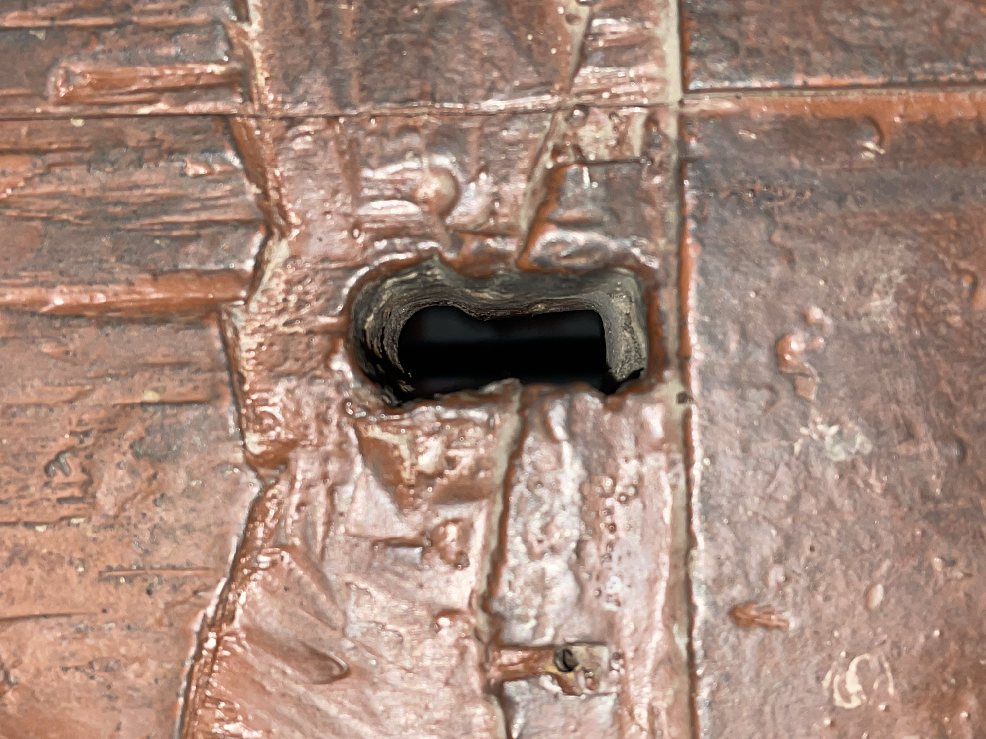 The image displays a close-up, high-detail view of a textured, reddish-brown surface, likely made of metal, stone, or ceramic, featuring a rough and uneven finish. The surface is segmented into irregular, raised rectangular patterns, resembling weathered bricks, tiles, or decorative panels. These segments are arranged both horizontally and vertically, creating a somewhat geometric yet rustic design.
A prominent, dark, and somewhat rectangular opening or hole is centered in the frame. The edges of this opening are irregular, suggesting it was either cast into the material or has undergone wear. The interior of the hole is deep black, indicating a hollow space or passage. The texture around the opening appears more pronounced, with varying shades and a slightly glossy sheen in certain areas.
The surface exhibits a range of reddish-brown hues, with darker tones in the recesses and lighter, almost silvery or pale beige highlights on the raised portions, possibly due to light reflection or wear. Small, irregular bumps, specks, and what appears to be dried liquid or residue are visible across the surface, contributing to its aged and weathered appearance. No individuals or specific activities are depicted. There is no visible text, and the image does not provide direct visual cues for time of day, weather, or specific location within Santa Ana, Venezuela, beyond the inherent characteristics of the material shown.