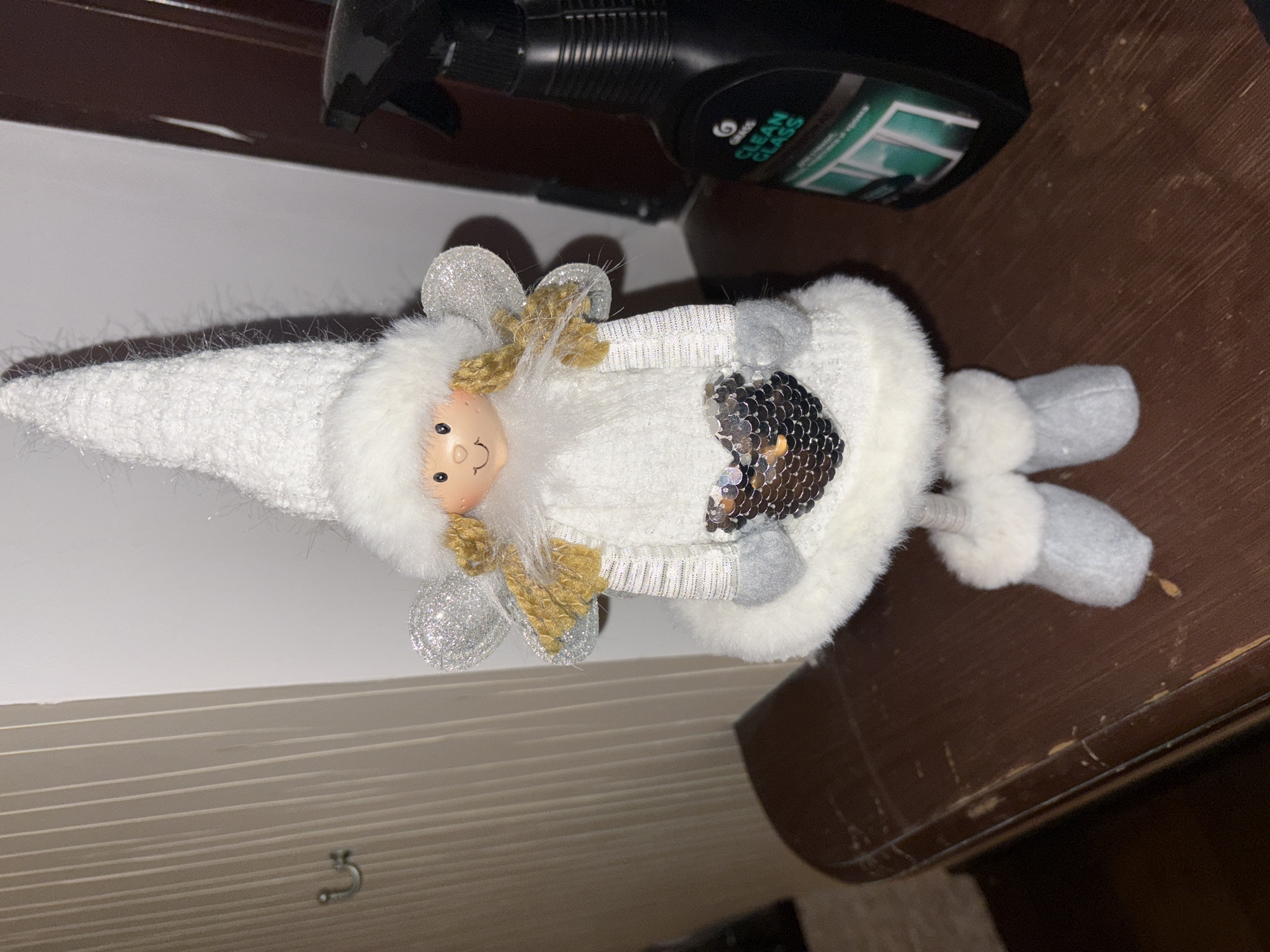 A slightly angled, close-up shot shows a white and silver Christmas angel doll lying on a dark brown wooden surface, possibly a table or shelf. The doll has a white pointed hat, a small painted face, and silver and gold wings. It has a sequined silver heart on its body and gray legs. A black spray bottle with a green label, likely for cleaning glass, is partially visible in the background. The location is unknown, and there is no information to identify the image as being in Ореховка, Russia.