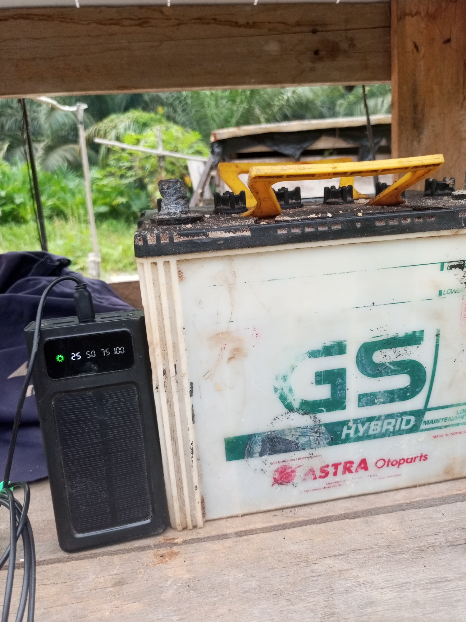 The image depicts a close-up, eye-level view of a car battery and a black power bank situated on a wooden surface, likely outdoors. The car battery, a GS Hybrid by Astra Otoparts, is white with green and red branding. It shows signs of wear and tear, with some dirt and corrosion around the terminals. The power bank, connected to the battery via a black cable, has a small digital display showing "25 50 75 100" and a green indicator light. A portion of a blue cloth or garment is visible behind the power bank. The background is blurred, but suggests a natural environment with greenery and palm trees, consistent with a rural or semi-rural setting in Keling Panggau, Indonesia. The overall impression is one of practical, functional use of electrical equipment in an outdoor, possibly remote, location.