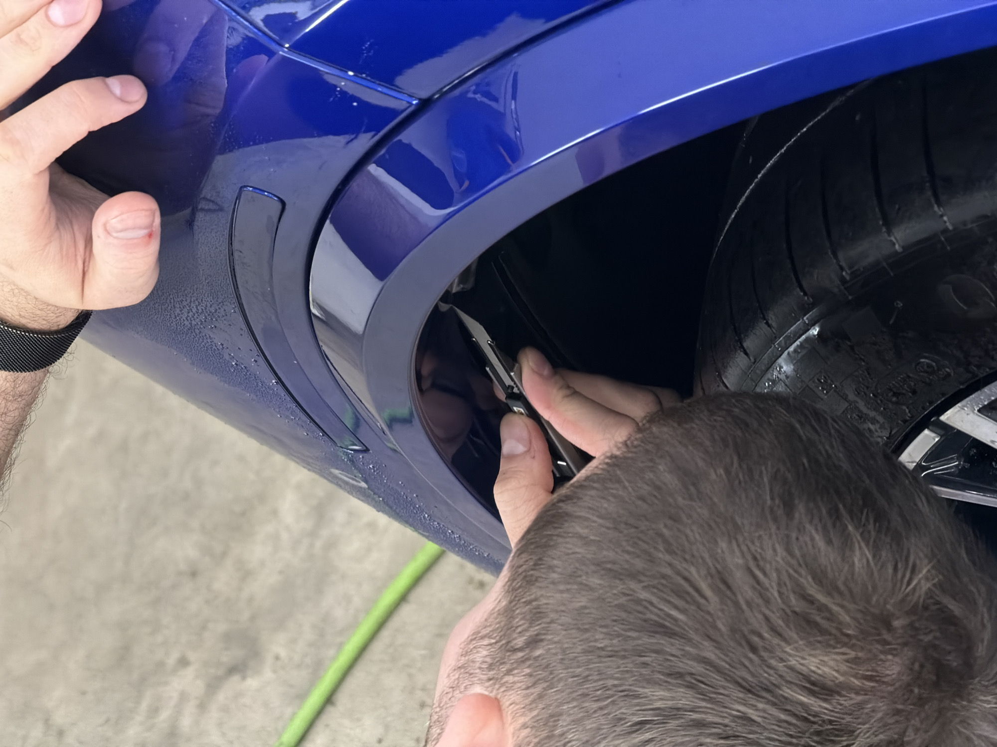 A person is working on the front quarter panel of a blue car, near the front tire. They are using a tool to manipulate something near the inner wheel well. The scene appears to be indoors, possibly a garage or shop, given the concrete floor and the presence of a green hose. The location cannot be determined from this image alone.