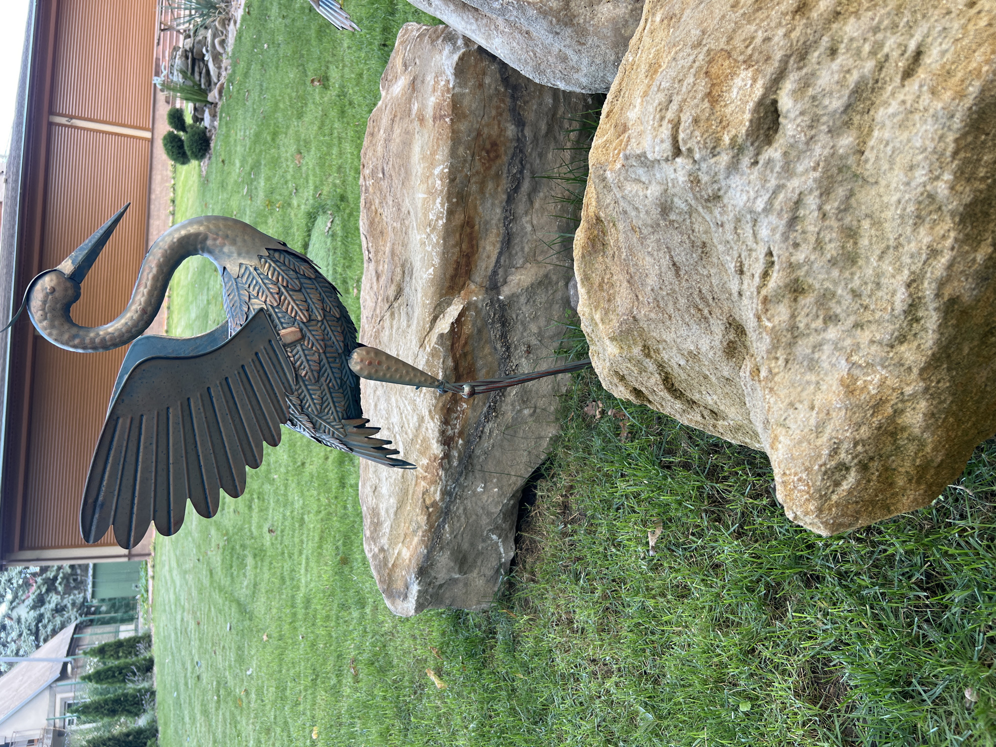 A metal sculpture of a heron stands on a large rock in a grassy yard. The heron is dark green and bronze, with its wings spread open. The yard is well-maintained, with neatly trimmed grass and a few fallen leaves. In the background, there is a house with a brown garage door and several evergreen trees. The scene is set in Kitskany, Moldova.