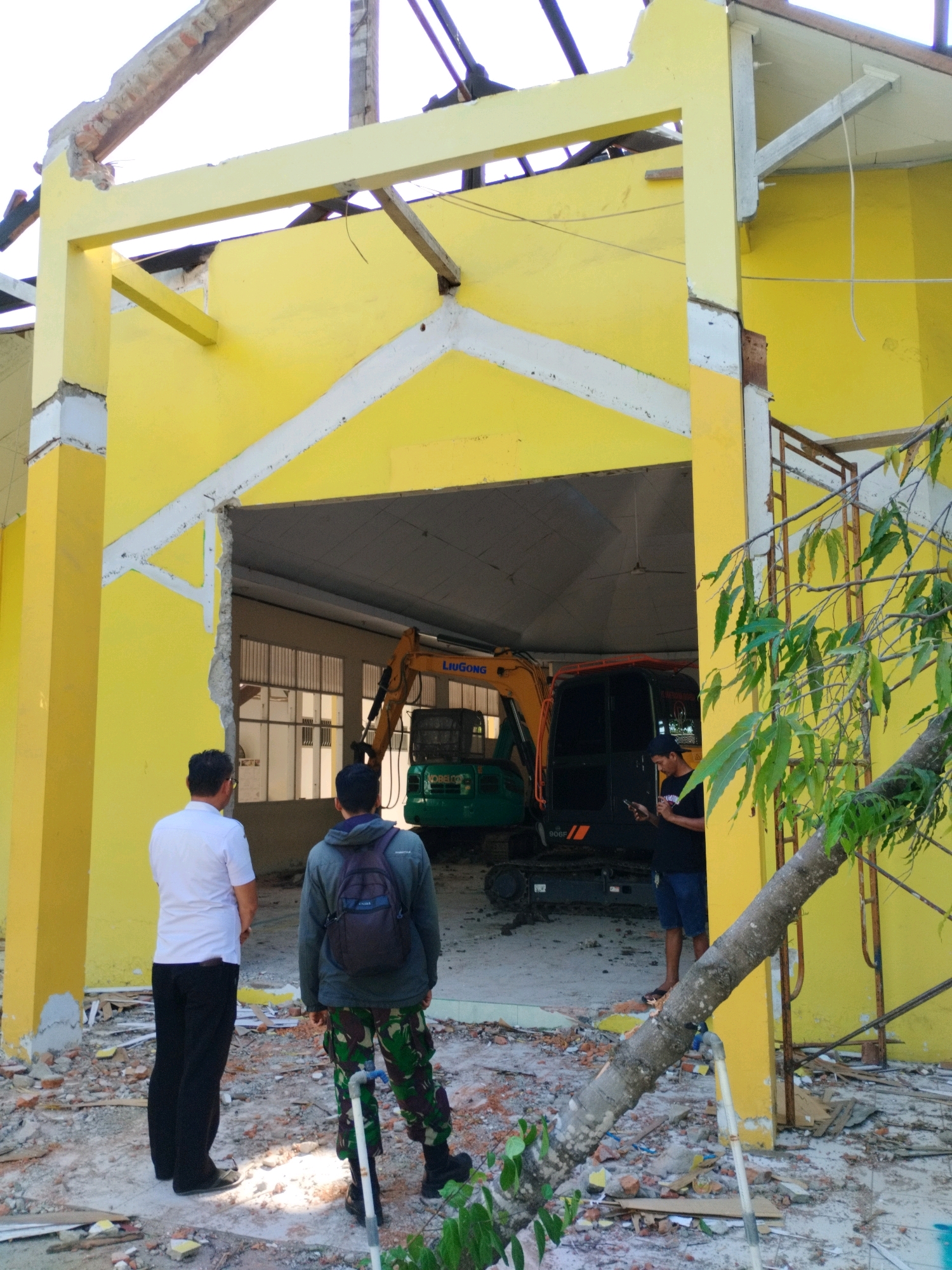 The image depicts a scene of demolition or significant structural damage to a yellow building. The roof appears to be partially collapsed, with exposed beams and debris scattered on the ground. An excavator, identified with the brand "LIUGONG", is visible inside the damaged structure. Three individuals are present: two men in the foreground looking towards the excavator, one wearing a white shirt and black pants, and the other in a camouflage-patterned uniform with a backpack. A third man, wearing a black t-shirt and blue shorts, is seen looking at his phone near the excavator. The environment suggests an outdoor setting, with evidence of construction or destruction materials like bricks and rubble. The image is taken in Gorontalo City, Indonesia.