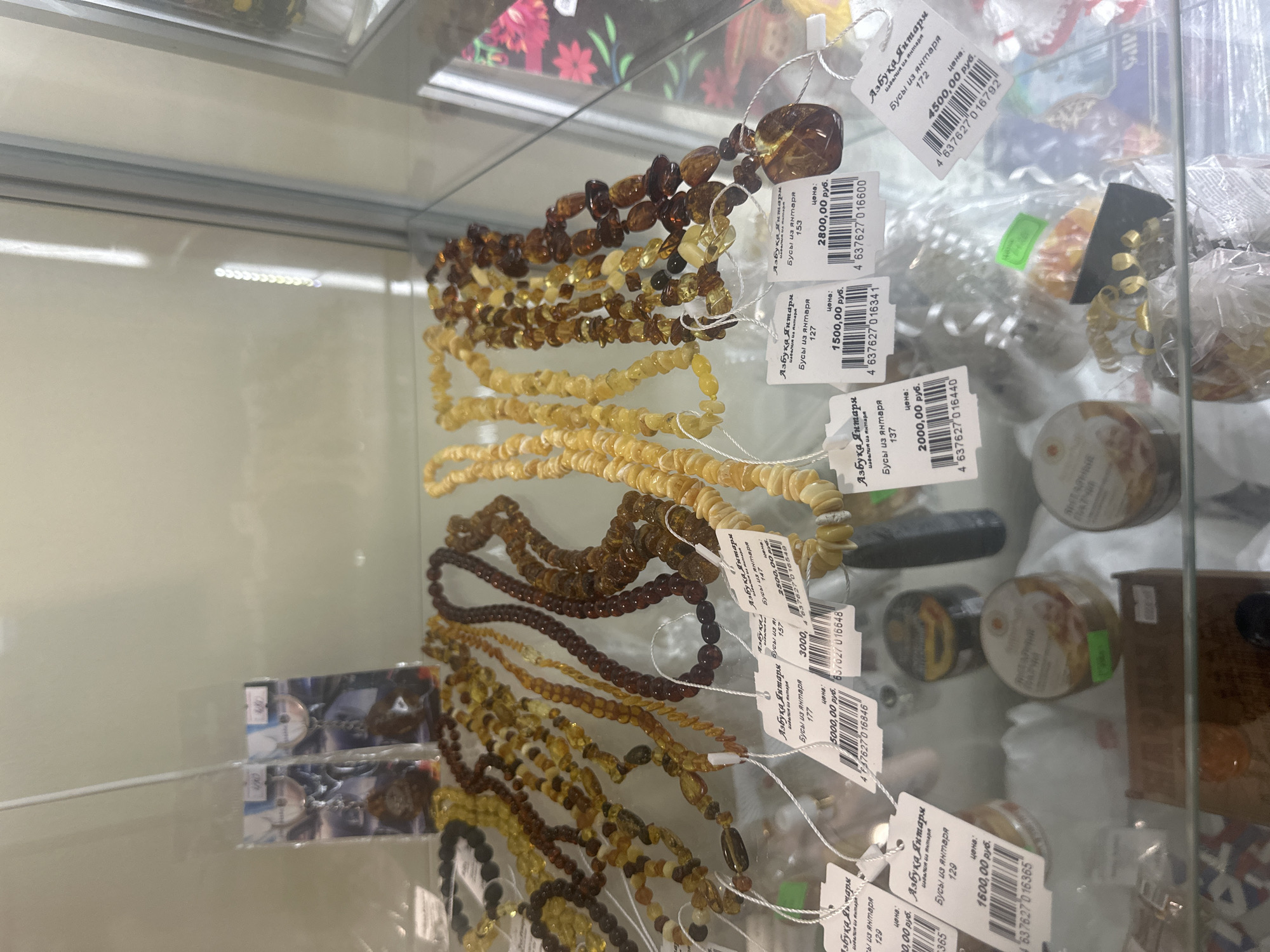 This media file captures a close-up view of several necklaces made of amber beads, displayed in a glass cabinet. The necklaces vary in color, ranging from light yellow to deep brown, and are arranged in cascading layers. Each necklace has a small tag attached, displaying Cyrillic text and a barcode. The text on the tags appears to indicate the product type ("Бусы из янтаря" - amber beads), a price (e.g., "2800,00 руб." - 2800.00 rubles, "1500,00 руб." - 1500.00 rubles), and potentially a product code or model number. The overall setting is a retail environment, likely a jewelry store or market stall, with other products visible in the background, some of which are also in packaging. The lighting is bright, suggesting an indoor setting during daylight hours. The focus is clearly on the amber jewelry, highlighting its natural variations in color and form. The scene is static, with no people visible, emphasizing the merchandise. The location context provided is Lyambir', Russia.
