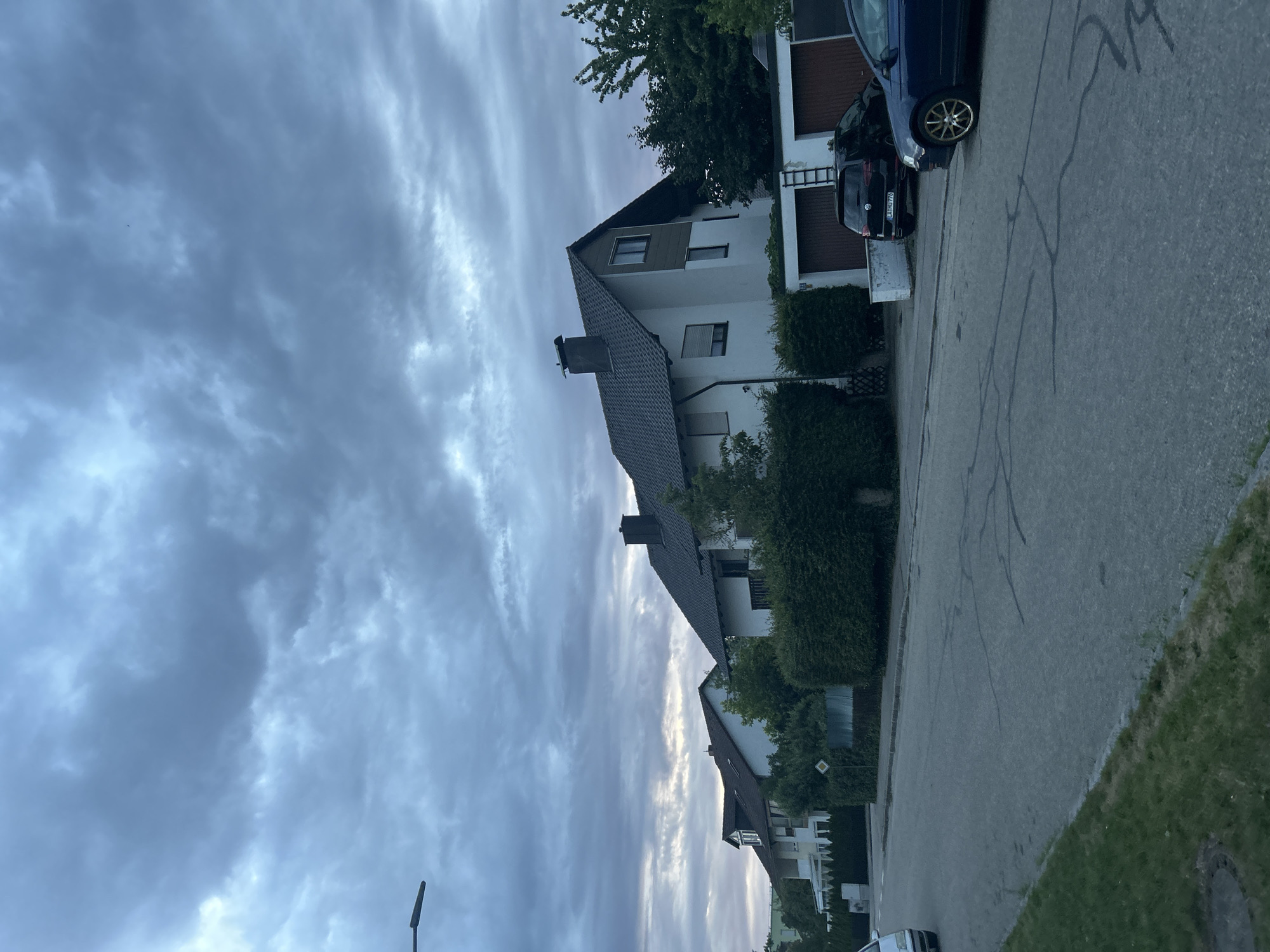 The image shows a suburban street in Ergolding, Germany, under a cloudy sky. Several houses line the right side of the street, characterized by pitched roofs and white facades, with well-maintained hedges in front of them. Two cars are parked on the street: a dark colored Volkswagen SUV and a blue sedan. The street itself is paved with asphalt and exhibits some wear and tear, with visible cracks. To the left of the street, a patch of grass and a sewer cover are visible. In the distance, more houses and trees are visible. A lamppost is present on the left side of the frame, extending into the upper left corner. The overall atmosphere is subdued due to the overcast conditions.