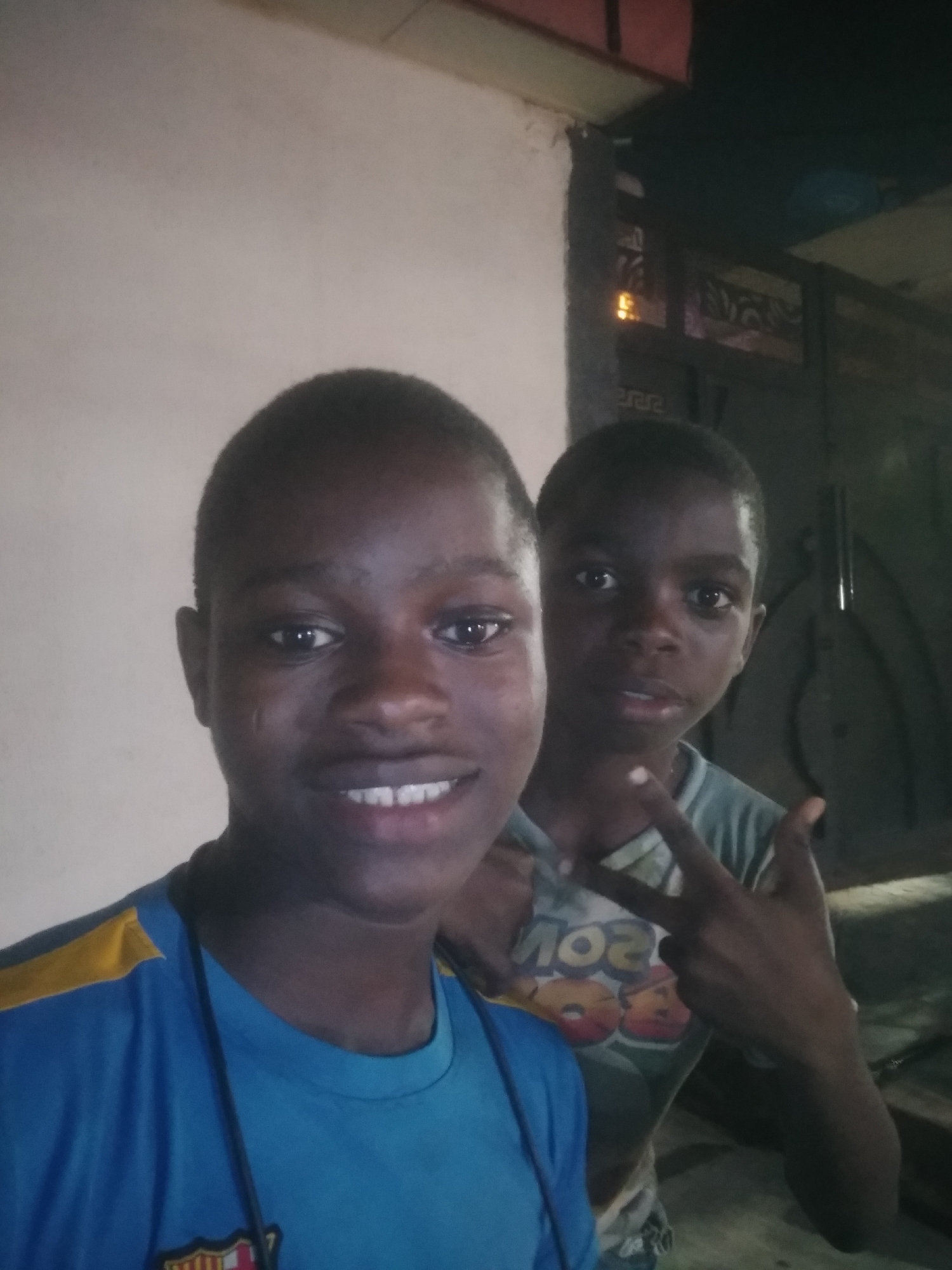 Two young boys are captured in what appears to be an evening or nighttime selfie, likely illuminated by a flash or nearby light source, in Shangisha, Nigeria. The boy in the foreground, a pre-teen or young teenager, has short dark hair and dark skin. He is smiling broadly, revealing his teeth, and looks directly at the camera. He wears a blue t-shirt with yellow shoulder panels and a partially visible dark crest or logo on the chest, along with a dark lanyard around his neck.
Behind him, slightly to the right, is a younger boy with dark skin and short dark hair. He is making a "peace sign" gesture with his right hand. His expression is more neutral or serious compared to the first boy. He wears a light-colored t-shirt with a graphic design that includes red and orange elements and visible letters, possibly "SOS" or "ZOS".
The background on the left consists of a plain, light-colored wall. On the right, the background is darker, featuring what appears to be a structural element like a pillar or door frame, alongside intricate dark metalwork, possibly part of a gate, window, or decorative screen. A warm, orange glow is visible through the metalwork, suggesting an interior or exterior light source in the vicinity. The overall setting appears to be a domestic or semi-public outdoor area at night.