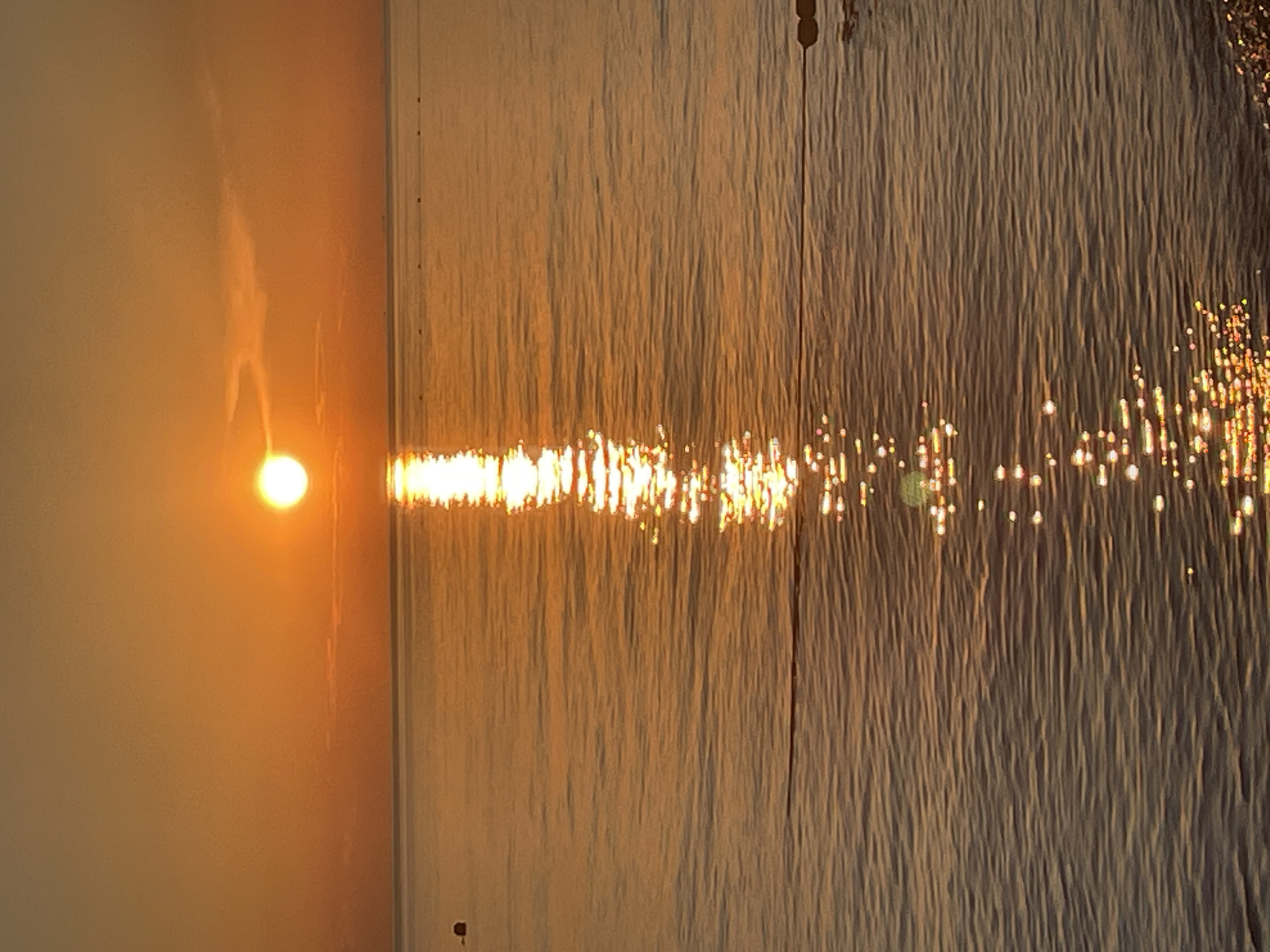 A serene sunset over a calm body of water is depicted. The sun, a bright orange orb, is positioned in the upper center of the frame, casting a long, shimmering reflection across the water's surface. The sky is a gradient of orange and yellow, with subtle cloud formations. The water itself is a warm, golden hue, with gentle ripples creating a textured appearance. A few small buoys are visible in the distance, marking the boundary of the water. A thin rope runs horizontally across the lower portion of the image, with a few buoys attached. The overall scene evokes a sense of tranquility and natural beauty.