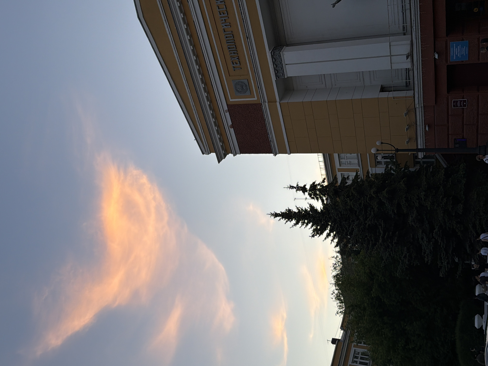 The image depicts the main building of what was formerly the Siberian State Technological University, now part of Reshetnev Siberian State University of Science and Technology, located in Krasnoyarsk, Russia.
The scene is set at dusk, under a pale blue sky. A prominent, elongated cloud, illuminated in vibrant orange and pink hues by the setting sun, stretches diagonally across the upper left quadrant of the sky. Smaller, fainter pink clouds are visible in the upper center.
The right half of the image is dominated by the multi-story university building, characterized by a yellow/ochre facade and white classical architectural elements, including cornices, decorative trim, and large pilasters or columns on the right side. A section of the upper facade on the left features reddish-brown textured brickwork. The building bears white Cyrillic text, "ТЕХНОЛОГИЧЕСК" (part of "Technological"), and a circular Soviet-era emblem depicting a hammer and sickle within a wreath, both in relief below the text. The base of the building is a darker reddish-brown brick.
In the mid-ground, several tall, dark green coniferous trees (likely spruces) are silhouetted against the sky. A denser grouping of dark green deciduous trees and bushes occupies the lower-left and central-left, partially obscuring a smaller, yellow building with white-framed windows.
In the lower foreground, a black lamppost with three spherical white luminaires stands centrally. To the right of the lamppost, a rectangular blue sign with white text is affixed to the building's reddish-brown base. Approximately 7-10 individuals are visible in the lower foreground, mostly in shadow, appearing to be walking or standing. Their clothing and specific actions are indistinct due to the low light and distance.