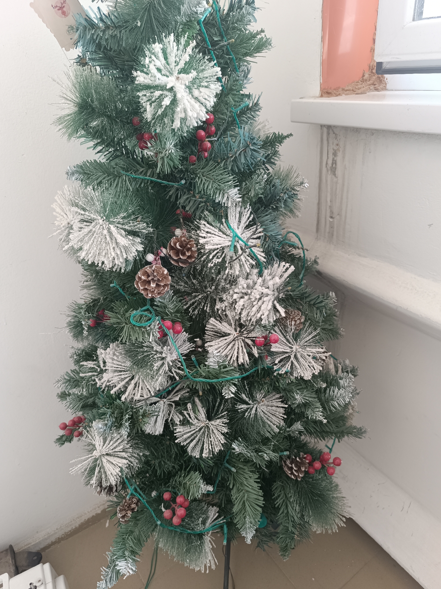 A close-up, vertical shot captures a decorated artificial Christmas tree in a room. The tree is adorned with green string lights, red berries, pinecones, and white, snowflake-shaped ornaments dusted with faux snow. The tree's branches are a dark green, appearing frosted with white. To the right of the tree, a white windowsill is visible, with a portion of an orange pipe and textured wall behind it. The floor below the tree appears to be tiled. The background is primarily a plain white wall. No people are present in the image. The scene suggests an indoor setting, possibly a home or an office decorated for the holidays. There is no information in the image to suggest the location is in the city of Taraz city administration, Kazakhstan.