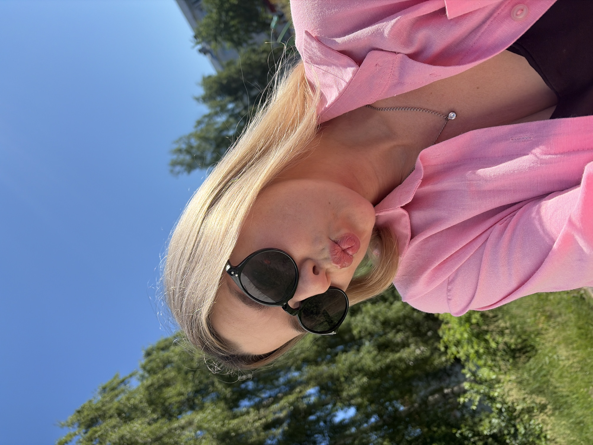 A young woman with blonde hair and sunglasses puckers her lips. She is wearing a pink button-up shirt and a silver necklace with a small pendant. The background is a blur of green trees and a bright blue sky, suggesting an outdoor setting. The presence of buildings in the background indicates an urban or suburban environment. The woman's pose and expression suggest she is taking a selfie.
