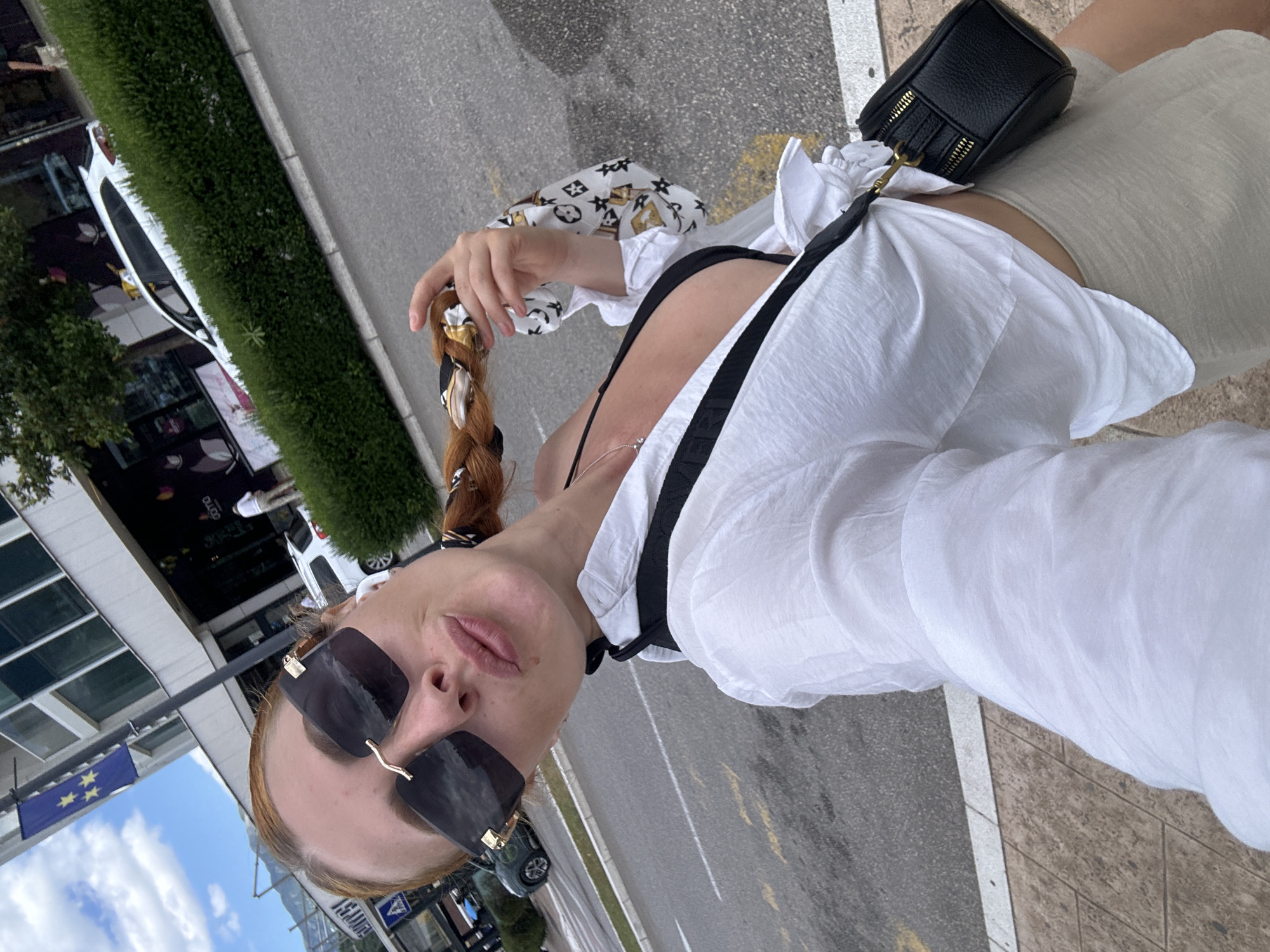 A woman with red hair styled in a braid, wearing large sunglasses, a black bikini top, an unbuttoned white shirt, and beige shorts, takes a selfie outdoors. She is holding a strand of her braided hair, which is adorned with a patterned scarf. A small black purse with gold zippers is slung across her body. She is standing on a paved sidewalk adjacent to a street with yellow lane markings. In the background, a building with a blue flag featuring stars is visible, along with a white car and a green hedge. The scene appears to be in the city of Budva, Montenegro, given the presence of the European flag and the architectural style of the building.