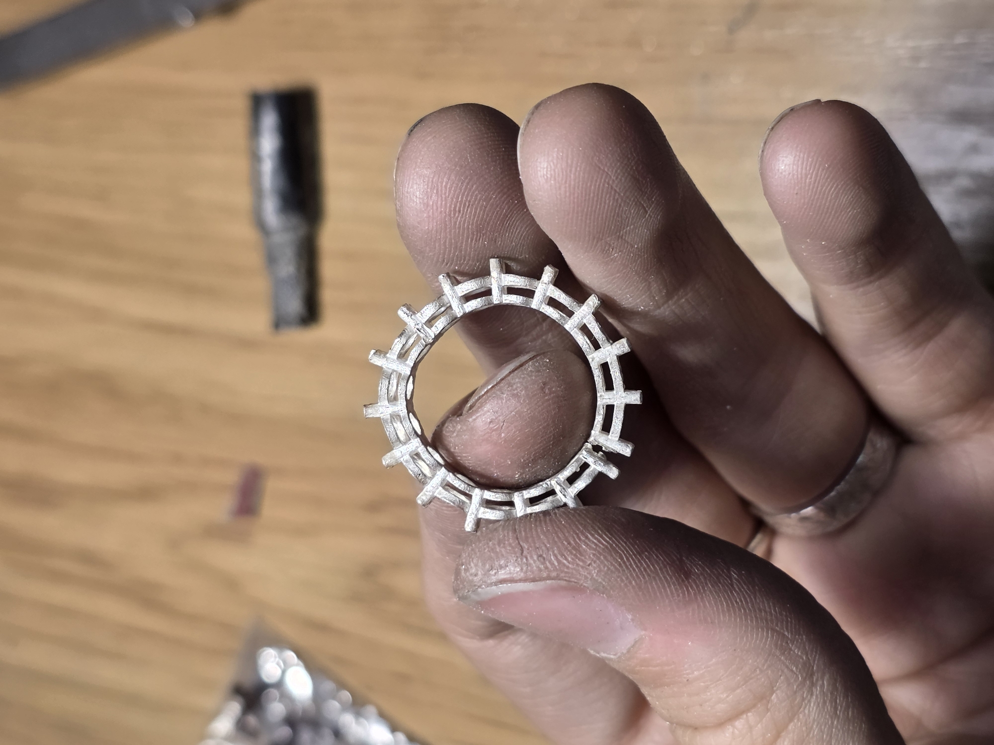 A person's hand holds a silver ring with a geometric design resembling a crown or gear. The ring is held in focus, with a wooden surface serving as the background. Tools and other small objects are blurred in the background, suggesting a workshop or crafting environment. No specific location within Kazan, Russia, can be determined from the image.