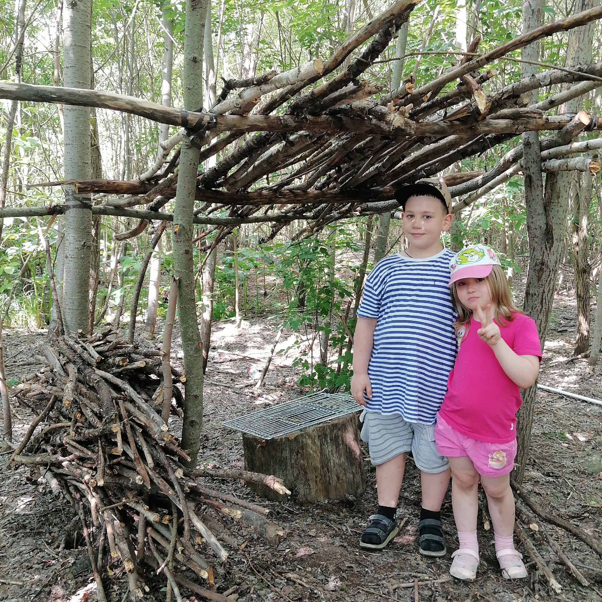 Two children, a boy and a girl, stand in a wooded area next to a rudimentary shelter constructed from branches. The boy, wearing a blue and white striped t-shirt, grey shorts, a black baseball cap, and black sandals, stands on the left. The girl, on the right, wears a bright pink t-shirt, pink shorts with a graphic print, a pink and white baseball cap, and light pink shoes with white socks. She is making a peace sign with her right hand. In the background, the shelter is made of numerous branches of varying sizes, creating a haphazard roof and walls. To the left of the children, a large pile of branches is stacked. A metal grill sits atop a tree stump to the right of the branch pile. The ground is covered in leaf litter and small branches. The environment appears to be a forest or park, with trees and foliage dominating the background. The lighting suggests it is daytime.